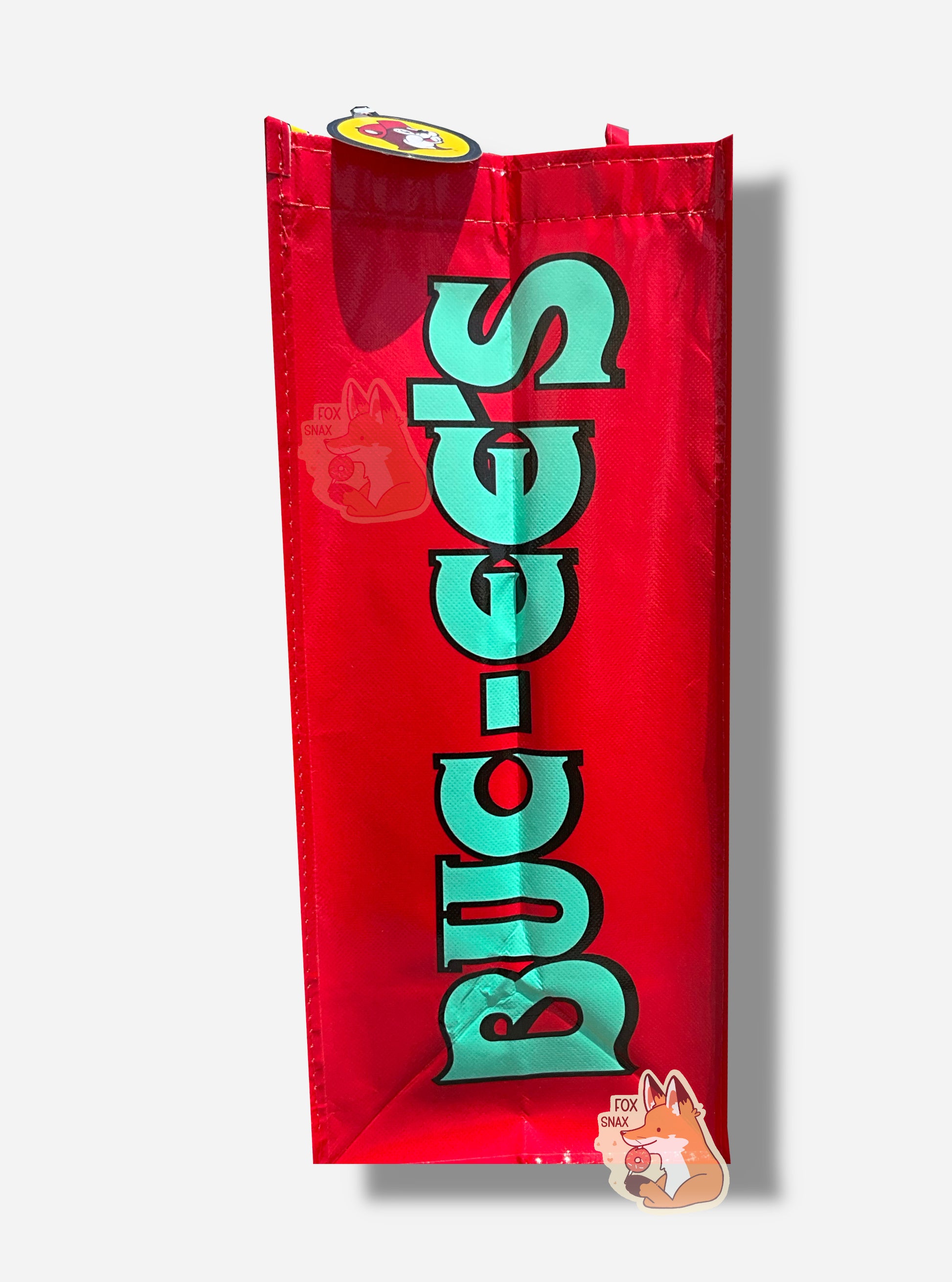 A picture of a the side of a large rectangular  plastic shopping bag.  The side of the bag has a red background, and reads BUC-EE'S in turquoise text with a black shadow.  Red straps can be seen poking out the top of the bag.