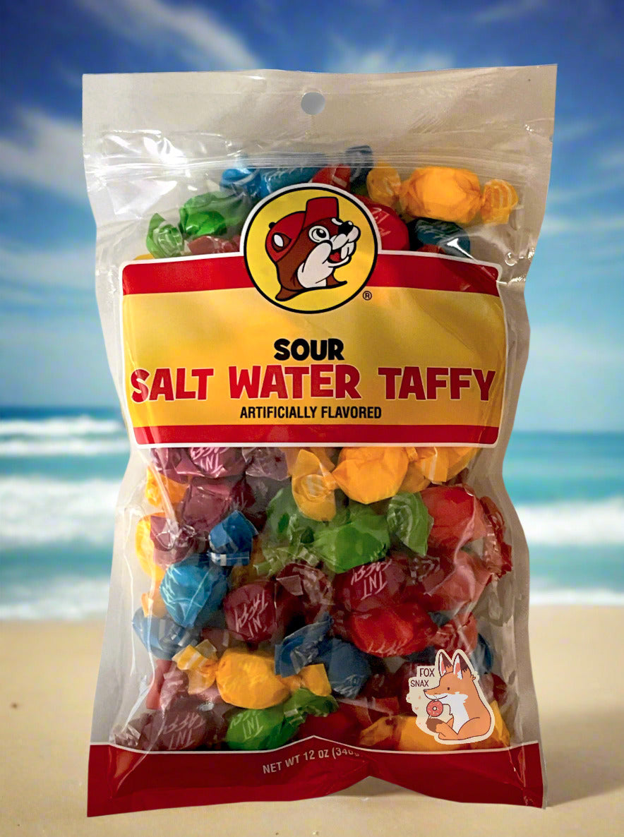 A picture of the front of a plastic bag of Buc-ee's Sour Salt Water Taffy. The bag is clear plastic with the Buc-ee's logo. Inside it, twists of candy in five different wrapping colors (red, yellow, green, purple) can be seen. The front of the bag has the Buc-ee's logo - a brown-furred beaver with a significant overbite, wearing a red ball cap - and reads: SOUR SALT WATER TAFFY, ARTIFICIALLY FLAVORED, then 12 OZ. The background is of a tan beach, with green/blue ocean waves, against a blue sky.