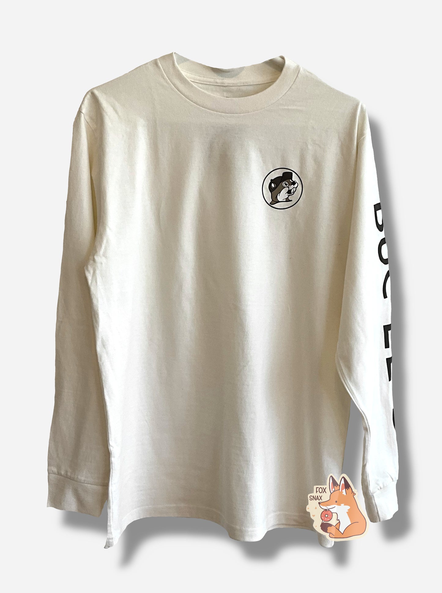 A picture of a white, long sleeve shirt, with the Buc-ee the Beaver logo on the right side (by left arm) on the chest. The logo is mostly muted out in grey and white hues, but Buc-ee's fur is a subtle brown. Running down the left arm, in large black text, reads BUC-EE'S.