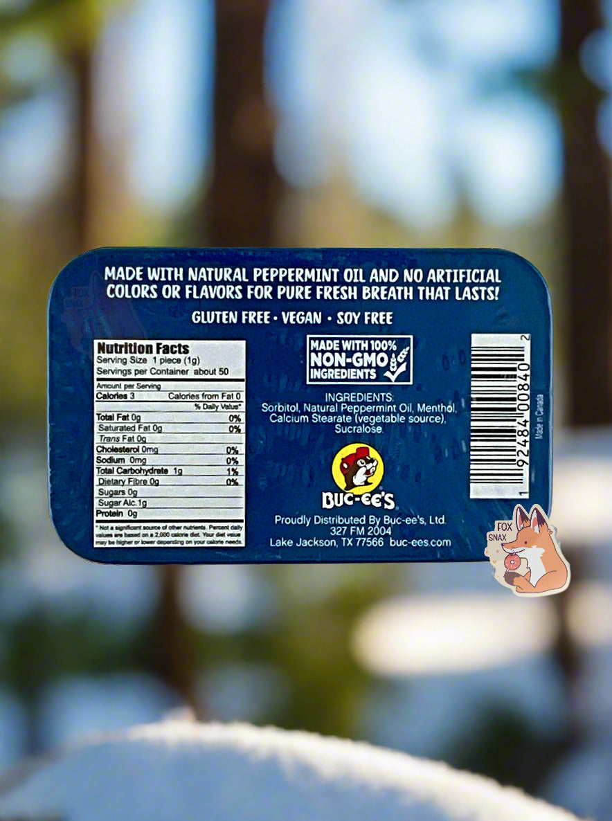 A picture of a back of a tin of Buc-ees Peppermint Mints. The back cover is decorated with a small picture of Buc-ee The Beaver, and has the following text: Made With Natural Peppermint Oil And No Artificial Colors Or Flavors For Pure Fresh Breath That Lasts!, Gluten Free, Vegan, Soy Free, made with 100% Non-GMO Ingredients. The background shows a snowy, forested winter wonderland, covered in pine trees.