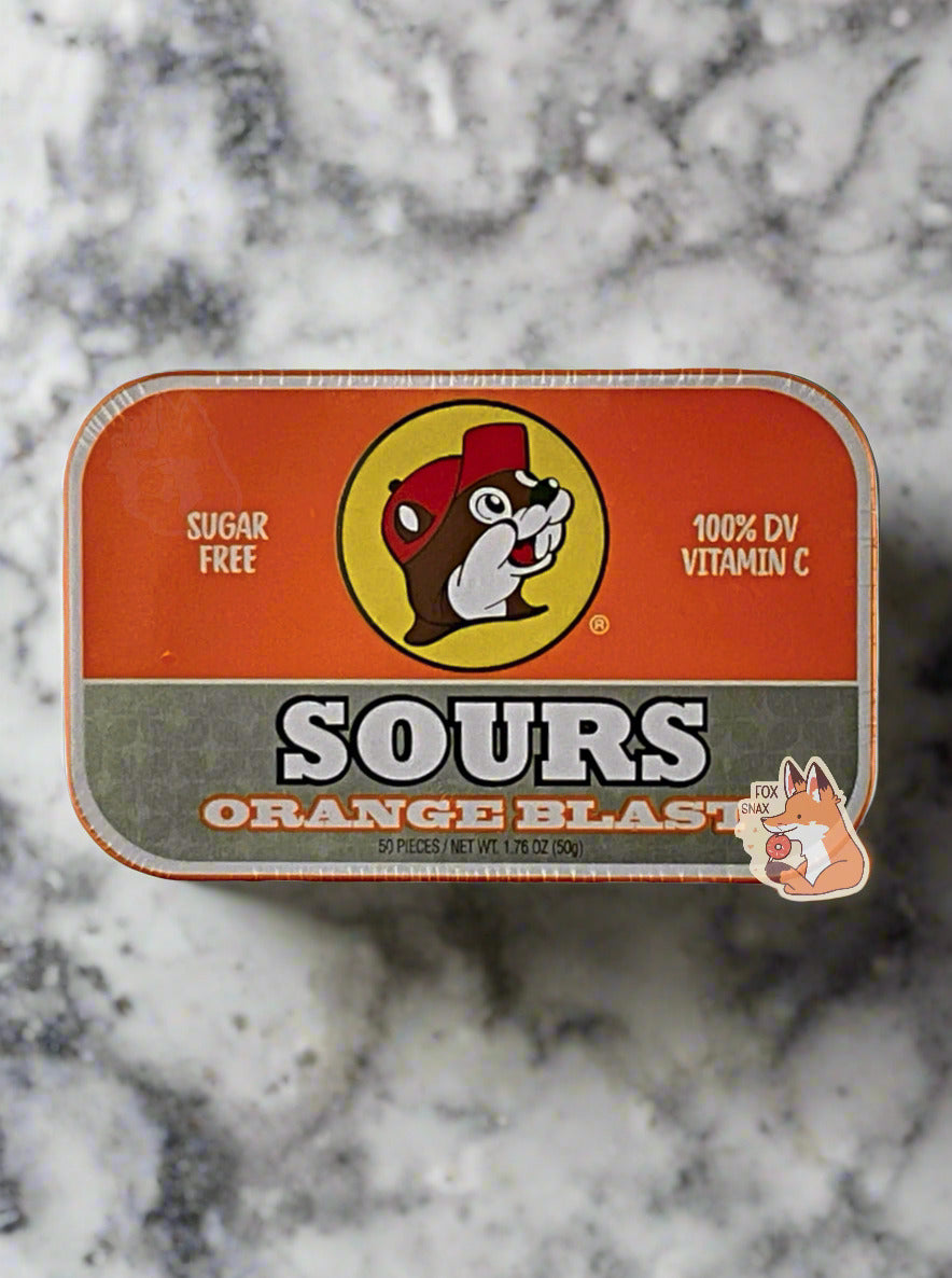 A picture of a tin of Buc-ees Orange Blast Sours. The cover is decorated with a big picture of Buc-ee The Beaver, and has an orange and silver background. The tin reads Sugar Free, 100% DV Vitamin C, Sours, Orange Blast, 50 pieces. The tin is sitting on a beautiful grey and black quartz countertop.