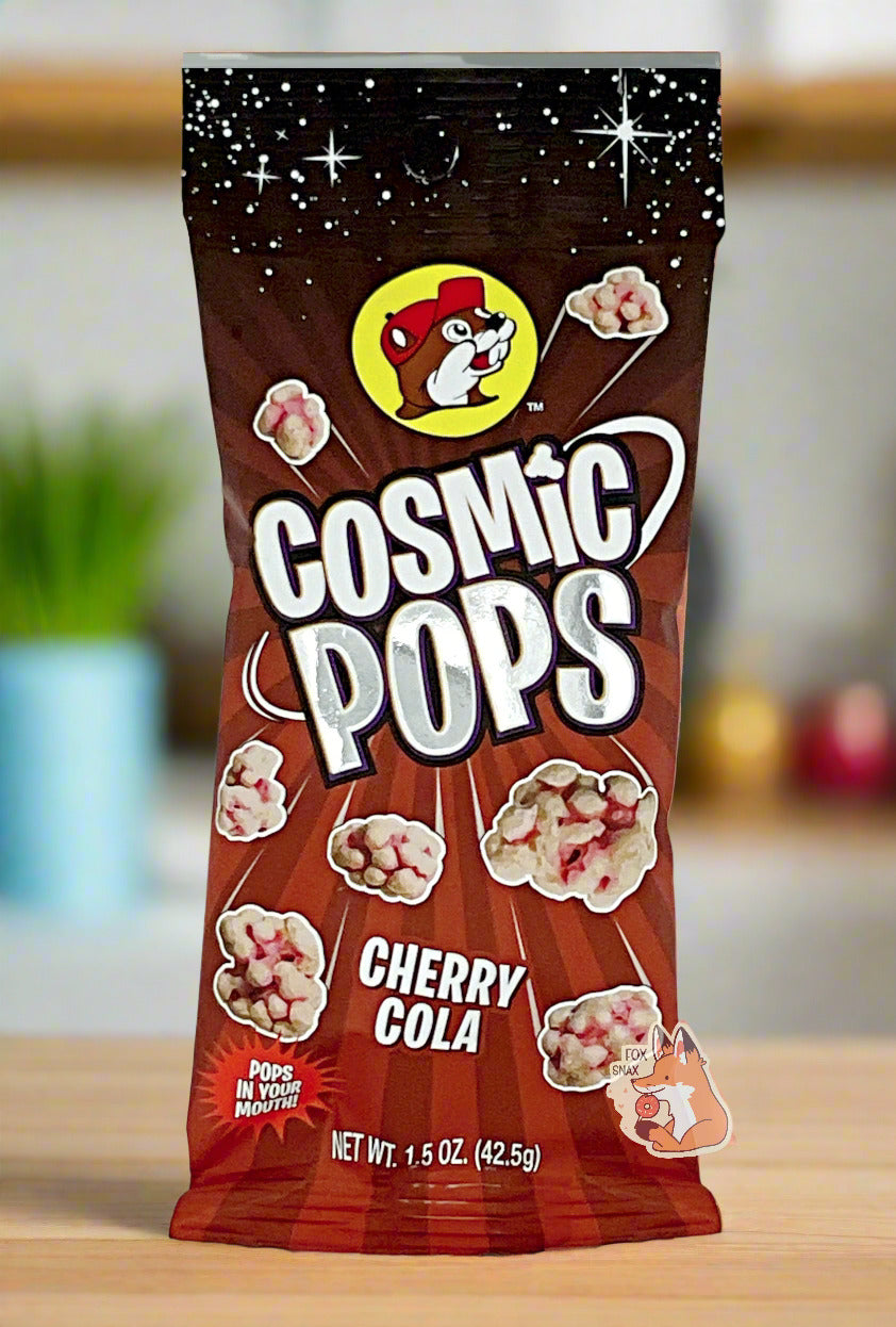 A picture of Buc-ee's Cosmic Pops, in Cherry Cola flavor. The bag starts with stars against a black background at the top, then shows rays of alternating red colors poking out of the center, to give the impression that the pops - lumpy odd shaped candies - are emerging from the center. A logo on the bottom reads POPS IN YOUR MOUTH! The background is of a bright cheery kitchen; the bag is resting on a wooden countertop.