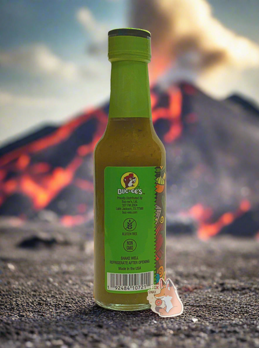 A picture of a small, tall glass bottle of hot sauce. The top of the bottle is sealed with a plastic top. The sauce inside is green/yellow with darker green flecks. In the background is an angry volcano, complete with lava.