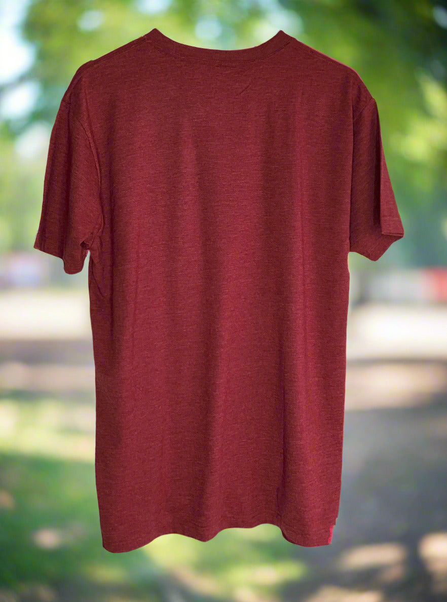 A picture of the back of a red t-shirt. The background is of a sidewalk lined with green trees and green grass, on a sunny day.