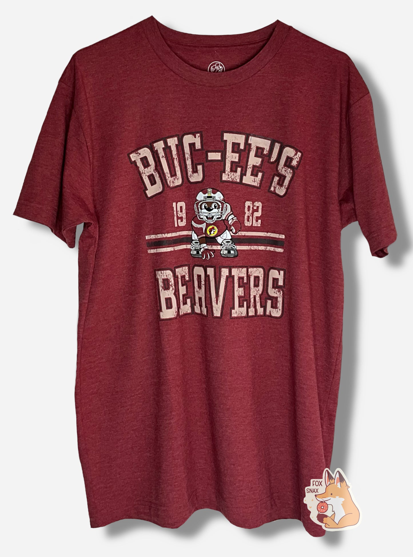 A picture of the front of a red t-shirt. The shirt has text on the top that reads BUC-EE'S, and text on the bottom reading BEAVERS. In the middle of the words are a picture of Buc-ee the Beaver wearing a dark red football uniform, complete with helmet. On either side of Buc-ee are the numbers 19 and 82.