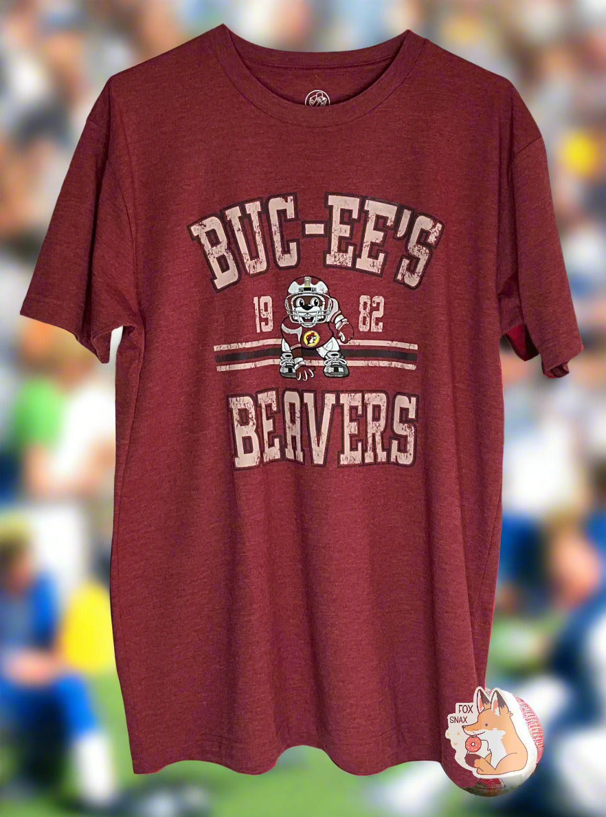 A picture of the front of a red t-shirt. The shirt has text on the top that reads BUC-EE'S, and text on the bottom reading BEAVERS. In the middle of the words are a picture of Buc-ee the Beaver wearing a dark red football uniform, complete with helmet. On either side of Buc-ee are the numbers 19 and 82. The background shows a football game's fans in the bleachers on a sunny day.