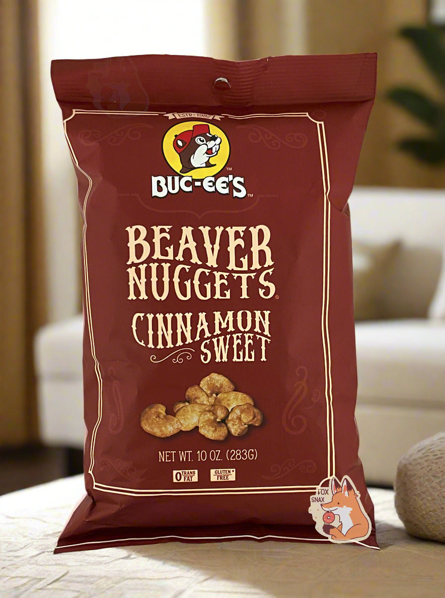 A picture of the front of a opaque, brown bag of Buc-ee's Beaver Nuggets, Cinnamon Sweet. which are puffed corn snack with cinnamon flavors, and has a picture of Buc-ee the Beaver on it. The bag reads: 0 Trans Fat. Gluten Free. The bag is sitting on a white couch in a clean living room; dark plants can be seen in the background.