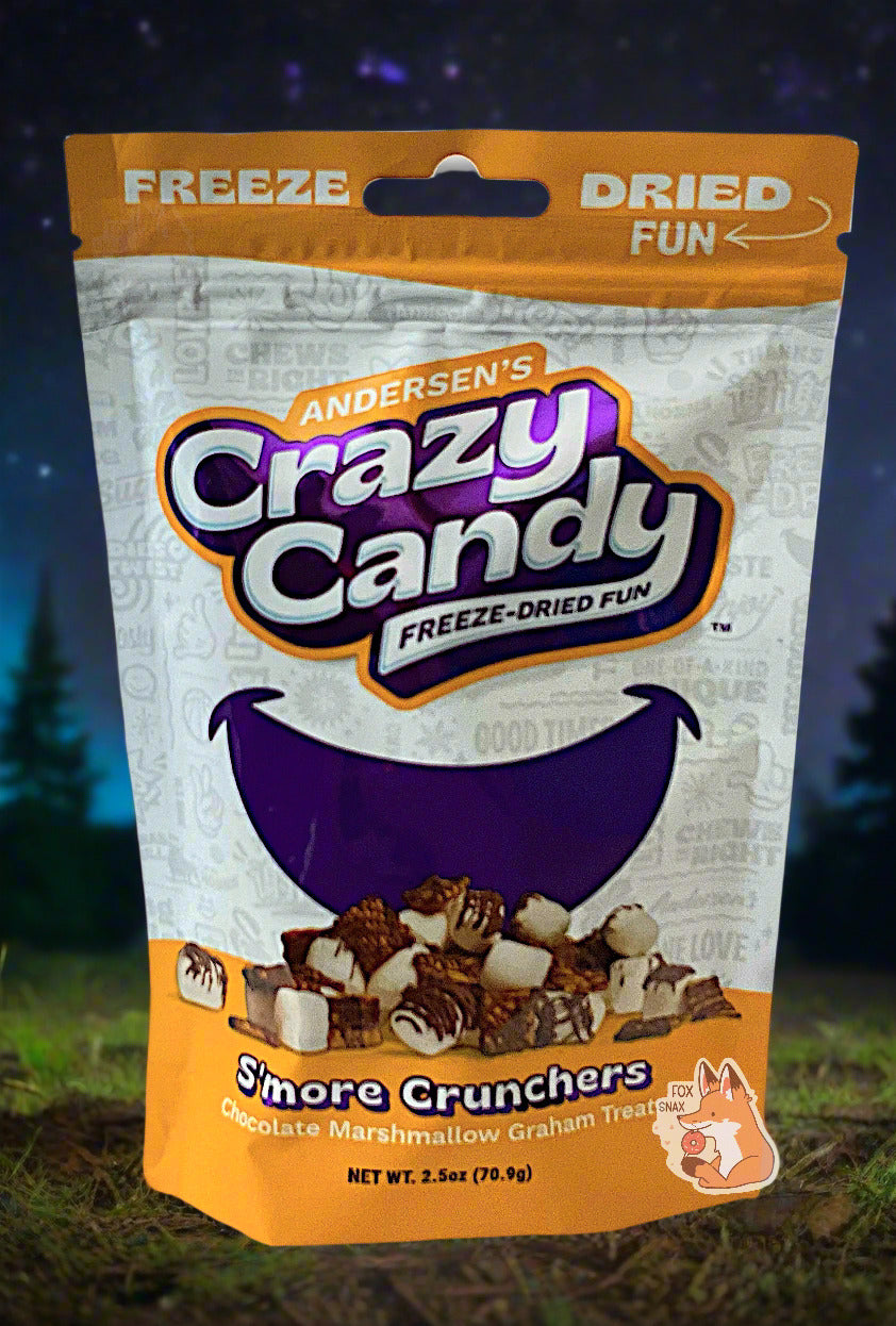 A large bag of Andersen's S'more Crunchers. The front of the bag also says Chocolate Marshmallow Graham Treats, Net Weight 2.5 oz. It's Fun, Happy, Candy. The top and bottom of the bag are yellow, and the background is a large purple smile against a white background. The background shows a campground; pine trees are illuminated against the night sky.
