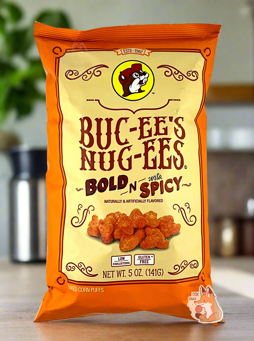 A picture of the front of a opaque, orange and yellow bag of Buc-ee's Nug-ee's Bold N Sorta Spicy, which are puffed corn snack with spicy flavors, and has a picture of Buc-ee the Beaver on it.  The bag reads: Naturally & Artificially Flavored.  Low Cholesterol.  Gluten Free.  Baked Corn Puffs.  The background is of a nice bright kitchen with green plants and kitchen accessories in the background.