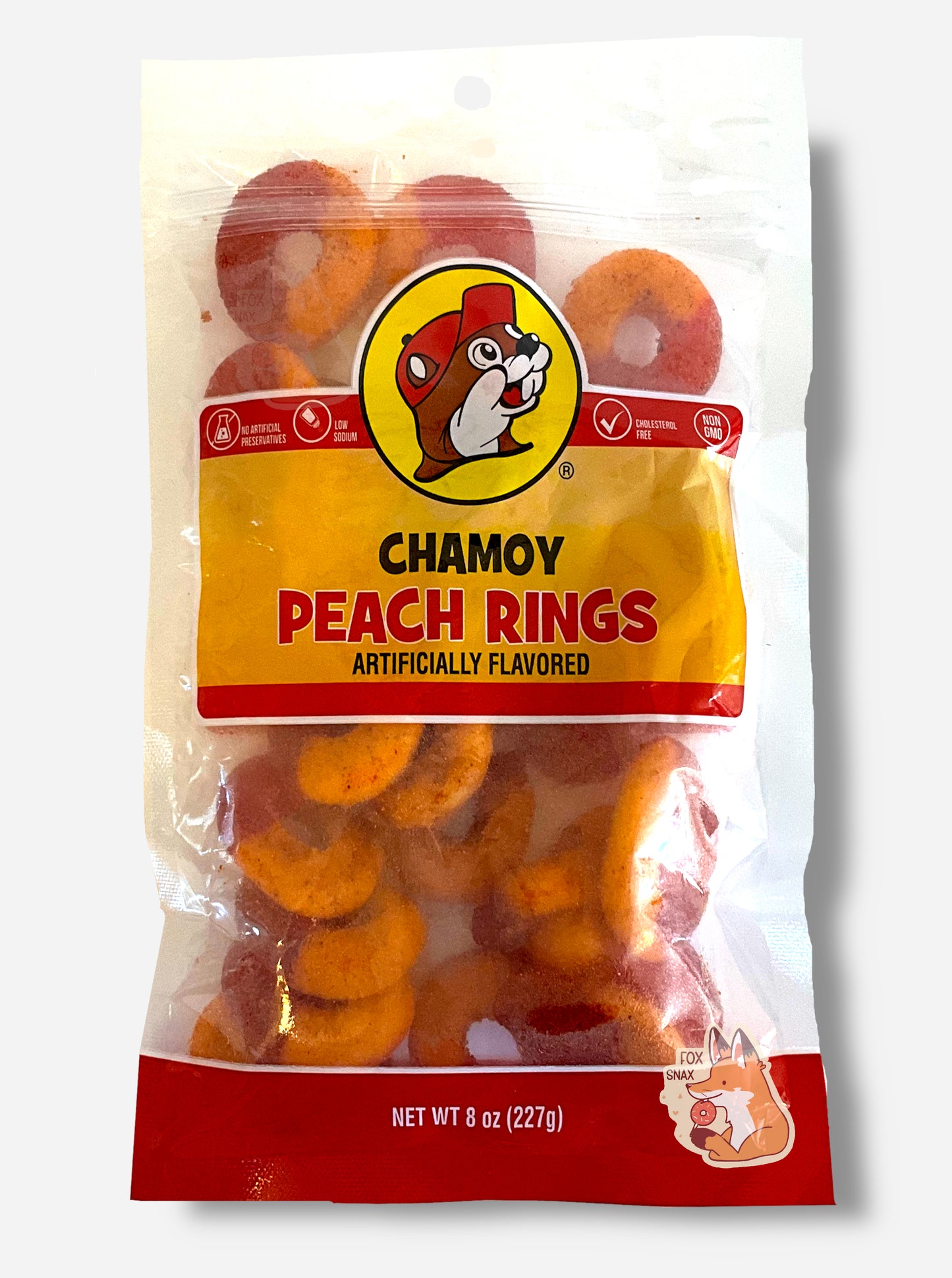 A picture of a clear plastic bag of Buc-ee's Chamoy Peach Rings. The bag reads ARTIFICIALLY FLAVORED, then NET WT 8 OZ (227 g), NO ARTIFICIAL PRESERVATIVES, LOW SODIUM, CHOLESTEROL FREE, then NON GMO. Inside the bag, peach rings can be seen; they are half red and half orange.