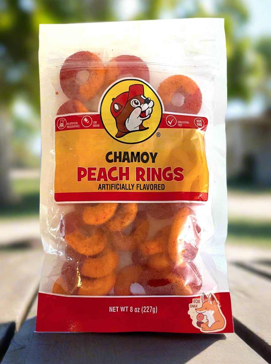 A picture of a clear plastic bag of Buc-ee's Chamoy Peach Rings. The bag reads ARTIFICIALLY FLAVORED, then NET WT 8 OZ (227 g), NO ARTIFICIAL PRESERVATIVES, LOW SODIUM, CHOLESTEROL FREE, then NON GMO. Inside the bag, peach rings can be seen; they are half red and half orange. The bag is sitting on an outdoor picnic table; peach trees can be seen in the background on a sunny day.