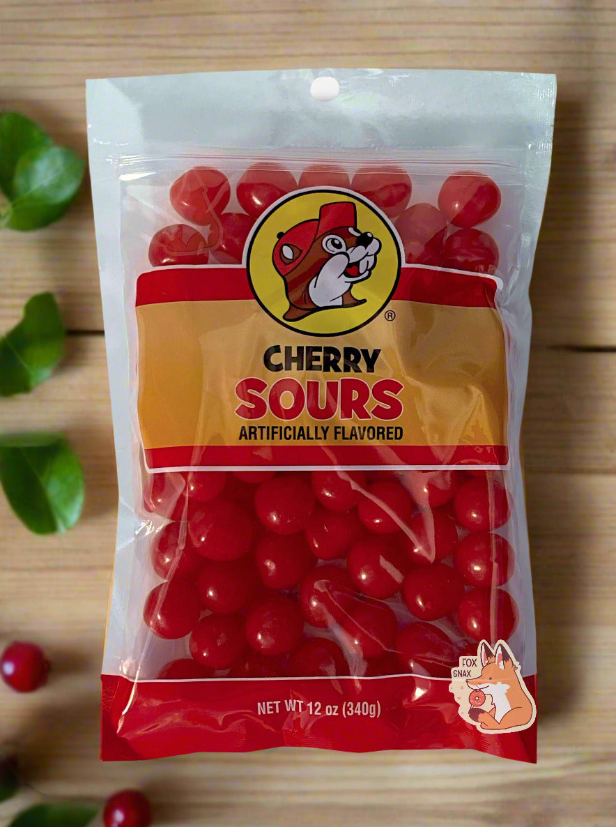 A picture of the front of a resealable bag of Buc-ee's Cherry Sours, which are red sour and sweet cherry candies. The bag reads CHERRY SOURS, Artificially Flavored, and has a picture of Buc-ee the Beaver on it. The sours inside are bright red and spherical. The background is of a light wood table, with leaves and cherries strewn about.