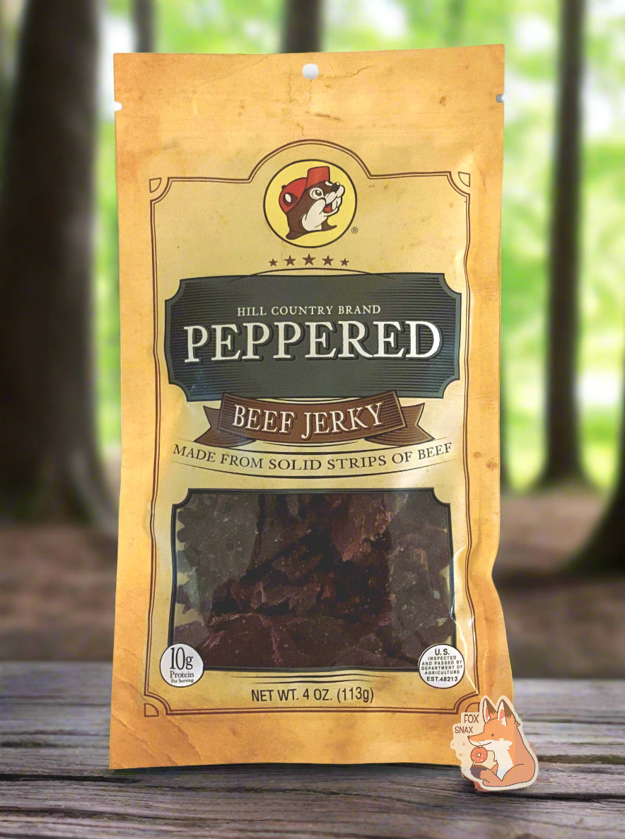 A picture of the front of a tan plastic bag of Buc-ee's Hill Country Brand Peppered Beef Jerky. The logo is in dark grey with white text. The label underneath reads 'Made From Solid Strips Of Beef'.A small window on the bottom of the bag shows the dark brown/red dried jerky inside. The bag has a picture of Buc-ee the Beaver on it. The bag is sitting on a rustic picnic table in a dense forest.