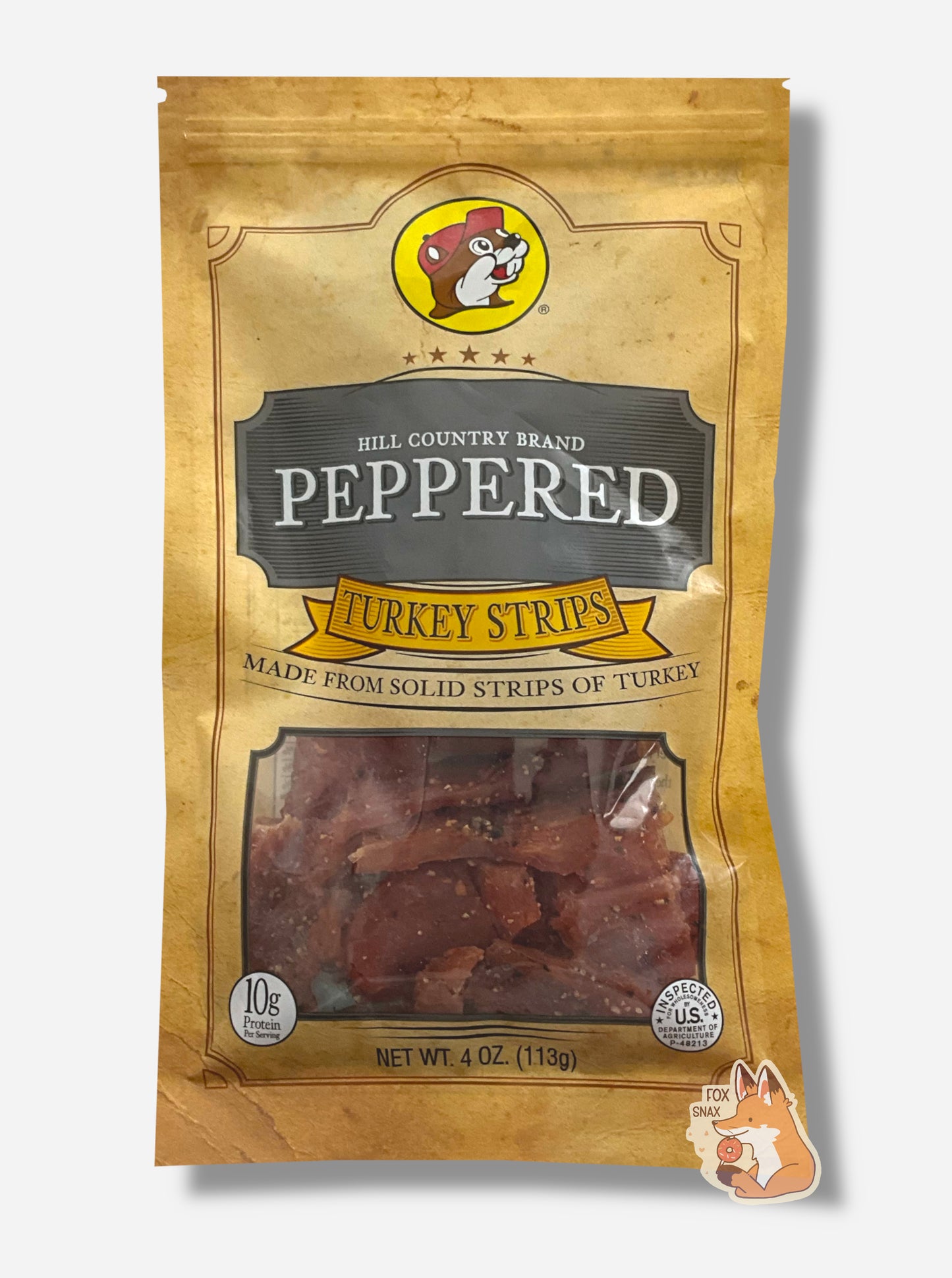 A picture of the front of a tan plastic bag of Buc-ee's Hill Country Brand Peppered Turkey Strips. The logo is in dark grey with white text. The label underneath reads 'Made From Solid Strips Of Turkey'.A small window on the bottom of the bag shows the dark red dried turkey jerky inside. The bag has a picture of Buc-ee the Beaver on it.