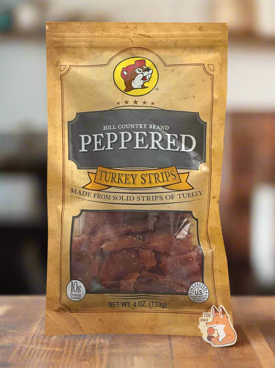 A picture of the front of a tan plastic bag of Buc-ee's Hill Country Brand Peppered Turkey Strips. The logo is in dark grey with white text. The label underneath reads 'Made From Solid Strips Of Turkey'.A small window on the bottom of the bag shows the dark red dried turkey jerky inside. The bag has a picture of Buc-ee the Beaver on it, and is sitting on a wooden table. In the background one can make out the vague details of a modern smokehouse.