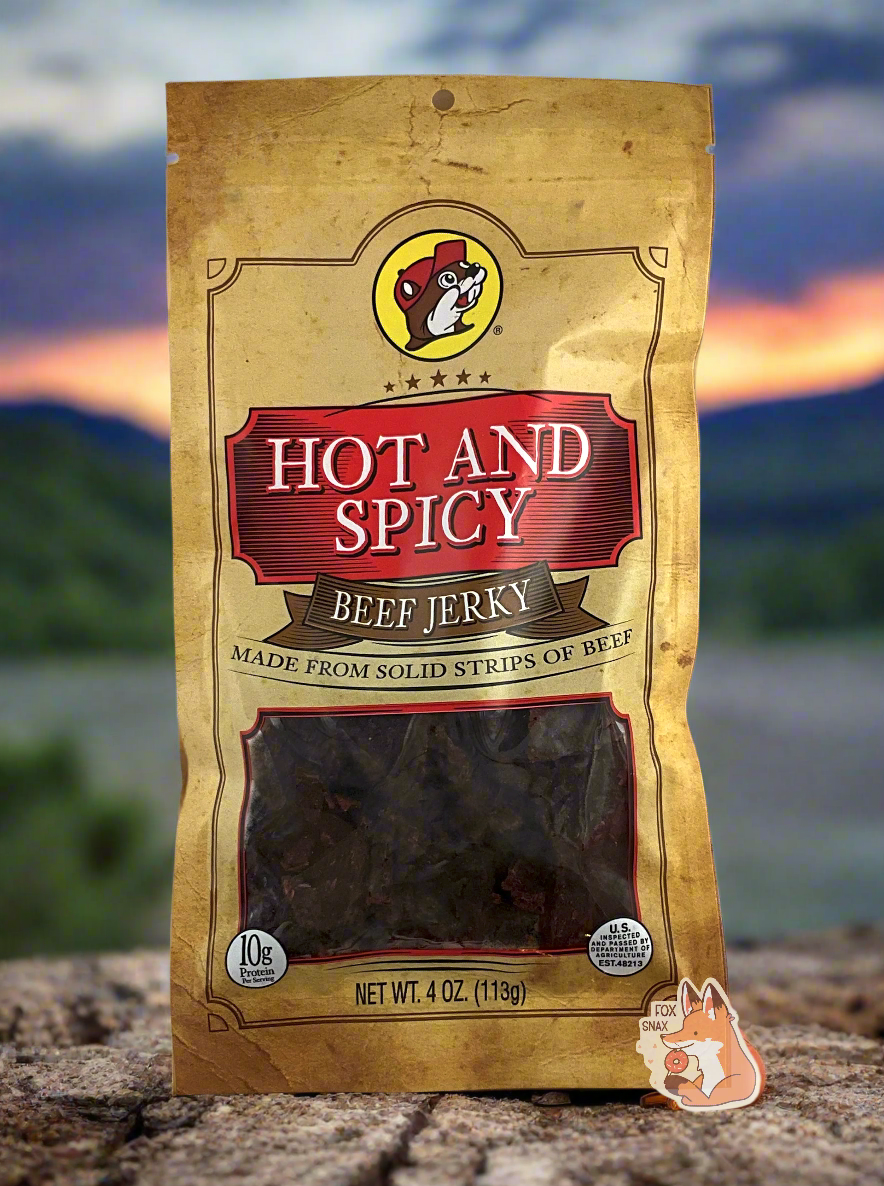 A picture of the front of a tan plastic bag of Buc-ee's Hot And Spicy Beef Jerky. The logo is in bright red with white text.The label underneath reads 'Made From Solid Strips Of Beef'.A small window on the bottom of the bag shows the dark brown/red dried jerky inside. The bag has a picture of Buc-ee the Beaver on it. The background shows campground with a beautiful sunset.