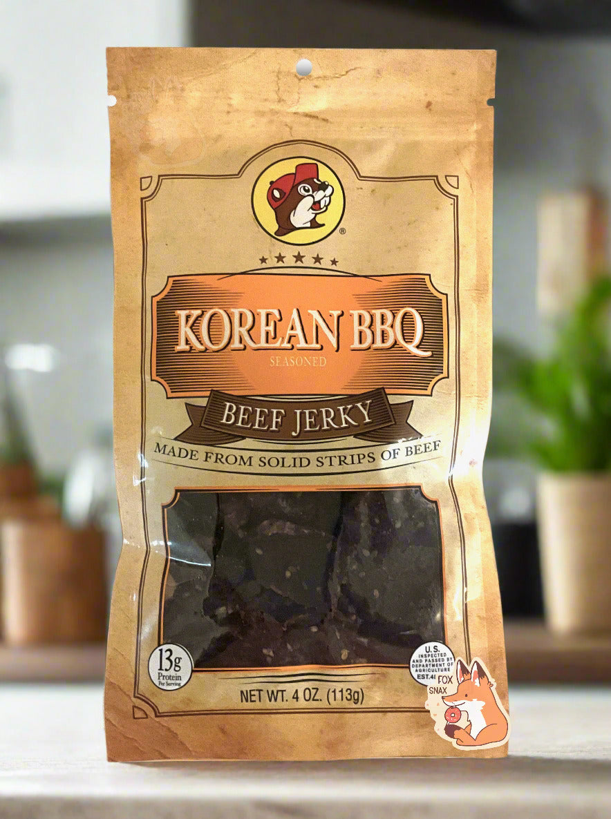 A picture of the front of a tan plastic bag of Buc-ee's Korean BBQ Beef Jerky.  The logo is in orange with white text.The label underneath reads 'Made From Solid Strips Of Beef'.A small window on the bottom of the bag shows the dark brown dried jerky inside.  The bag has a picture of Buc-ee the Beaver on it.  The background is a light, white-paneled kitchen with green plants in pots in the background.