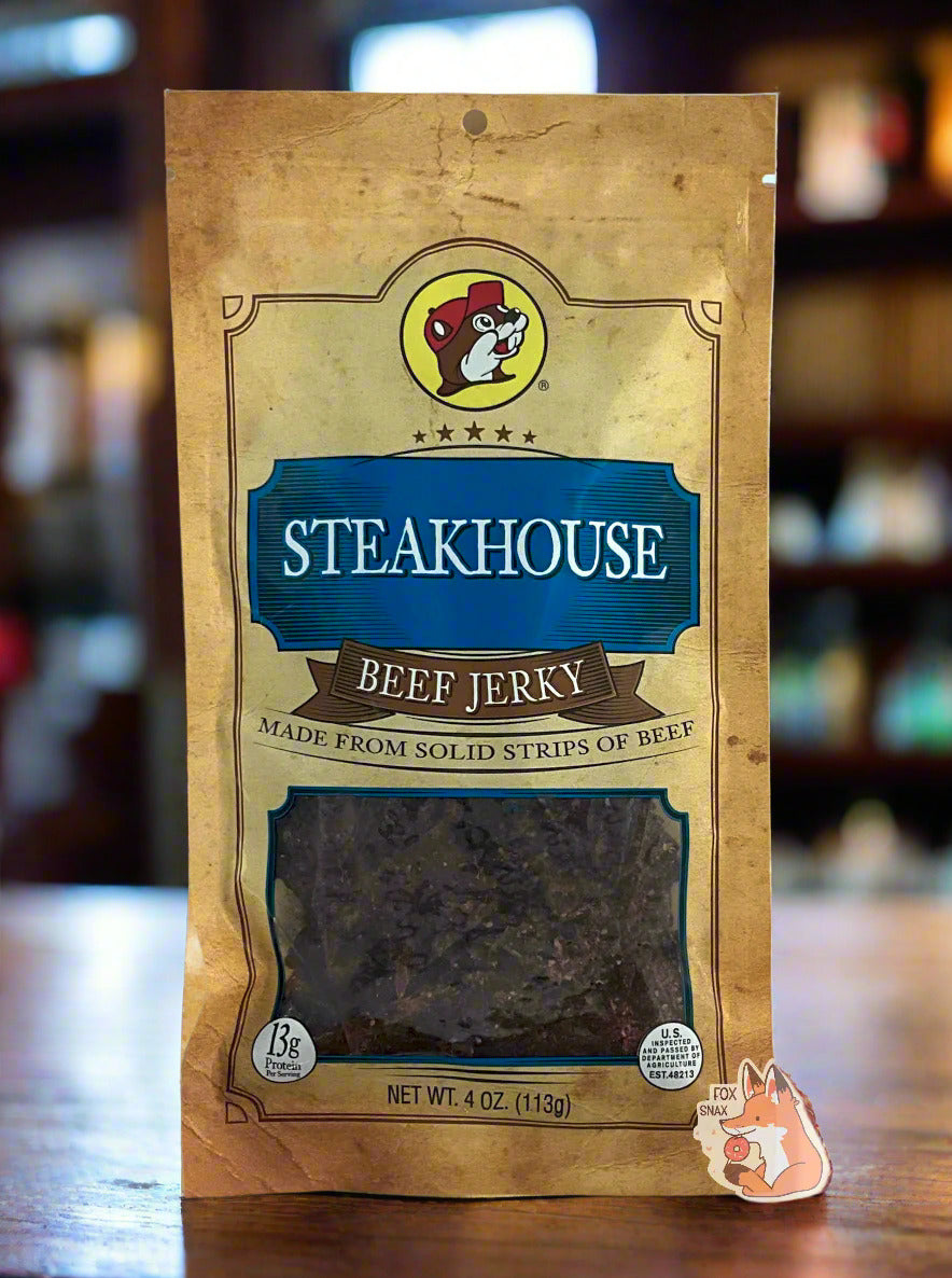 A picture of the front of a tan plastic bag of Buc-ee's Steakhouse Beef Jerky. The logo is in dark blue with white text. The label underneath reads 'Made From Solid Strips Of Beef'.A small window on the bottom of the bag shows the dark brown/red dried jerky inside. The bag has a picture of Buc-ee the Beaver on it. The bag is sitting on a wooden steakhouse table. The background shows the a steakhouse bar/restaurant with lots of dark wood.
