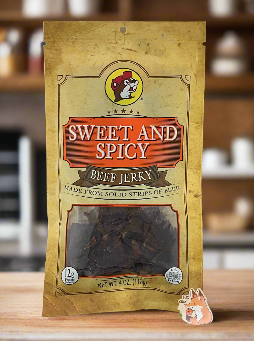 A picture of the front of a tan plastic bag of Buc-ee's Sweet And Spicy Beef Jerky. The logo is in orange with white text. The label underneath reads 'Made From Solid Strips Of Beef'.A small window on the bottom of the bag shows the dark brown/red dried jerky inside. The bag has a picture of Buc-ee the Beaver on it. The background is of a fancy, dark-wood paneled kitchen, with plants in the background. The bag of jerky is resting on a wooden table.
