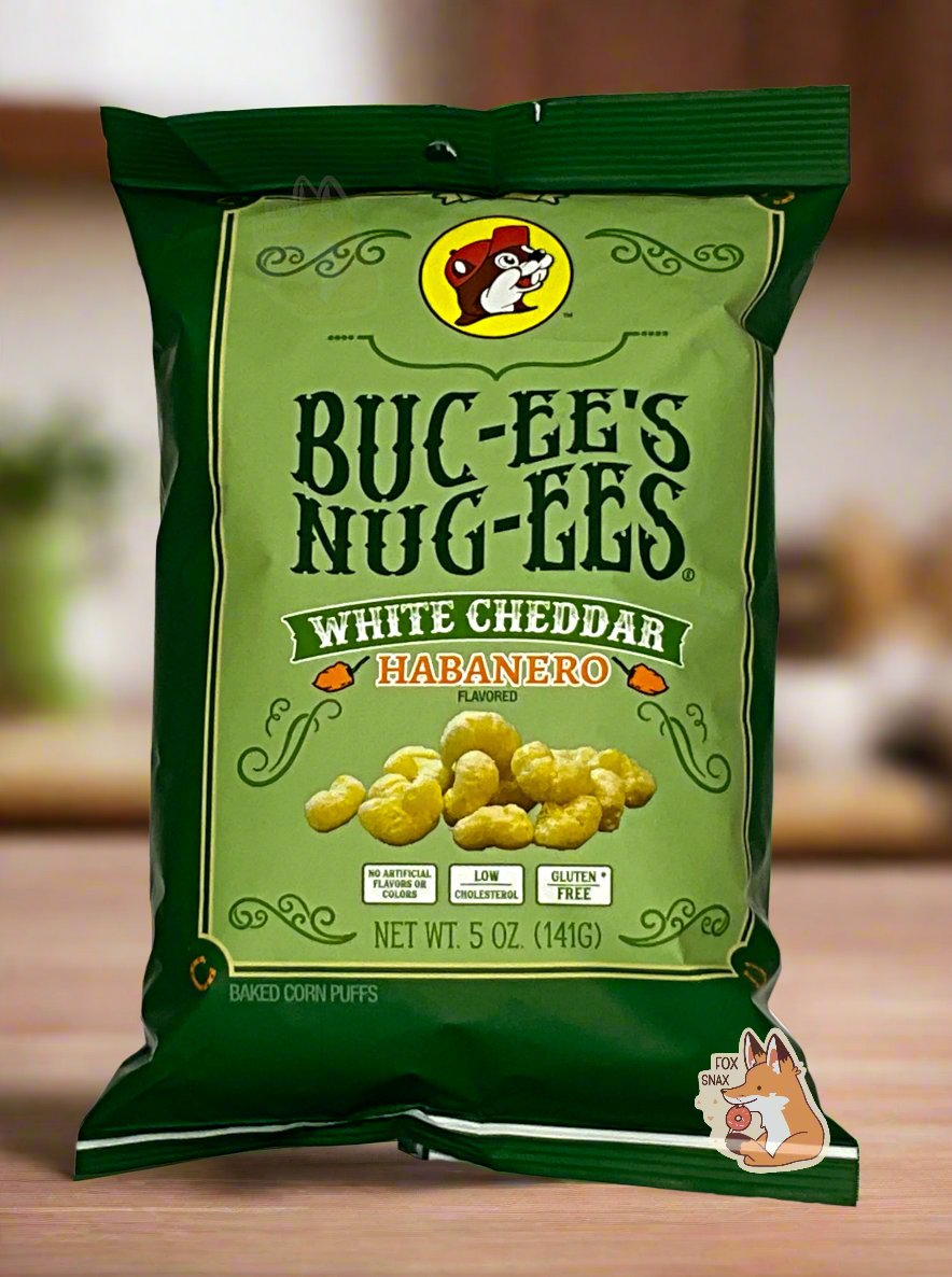 A picture of the front of a opaque, dark green and light green bag of Buc-ee's Nug-ees White Cheddar Habanero which are puffed corn snack with white cheddar and habanero pepper flavor, and has a picture of Buc-ee the Beaver on it. The bag reads White Cheddar Habanero Flavored, No Artificial Colors or Flavors, Low Cholesterol, Gluten Free, Baked Corn Puffs. The bag is sitting on a wooden kitchen countertop.