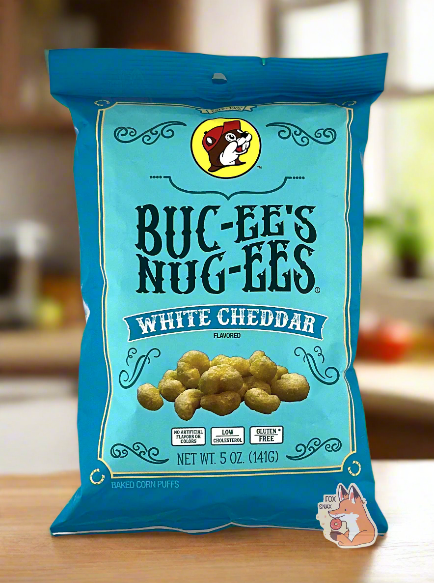 A picture of the front of a opaque, dark blue and light blue bag of Buc-ee's Nug-ees White Cheddar which are puffed corn snack with white cheddar flavor, and has a picture of Buc-ee the Beaver on it.  The bag reads White Cheddar Flavored, No Artificial Colors or Flavors, Low Cholesterol, Gluten Free, Baked Corn Puffs.  The bag is sitting on a wooden countertop in a nice, brightly lit kitchen with wooden cabinets.