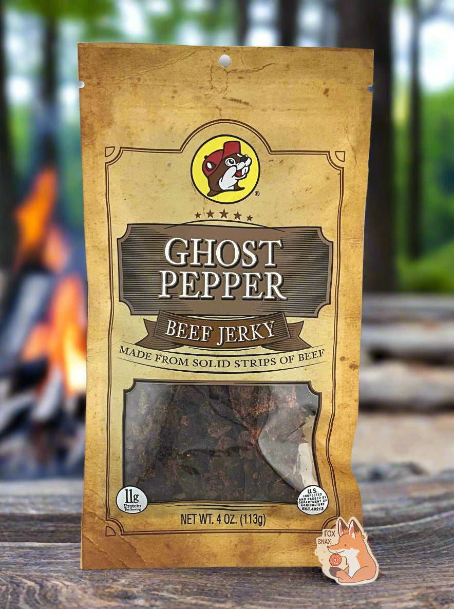 A picture of the front of a tan plastic bag of Buc-ee's Ghost Pepper Beef Jerky. The logo is in tan with white text. The label underneath reads 'Made From Solid Strips Of Beef'.A small window on the bottom of the bag shows the dark brown/red dried jerky inside. The bag has a picture of Buc-ee the Beaver on it, and is sitting on a piece of smooth wood. In the background, a giant campfire with open flames can be seen, and behind that, a green forest with tall trees.