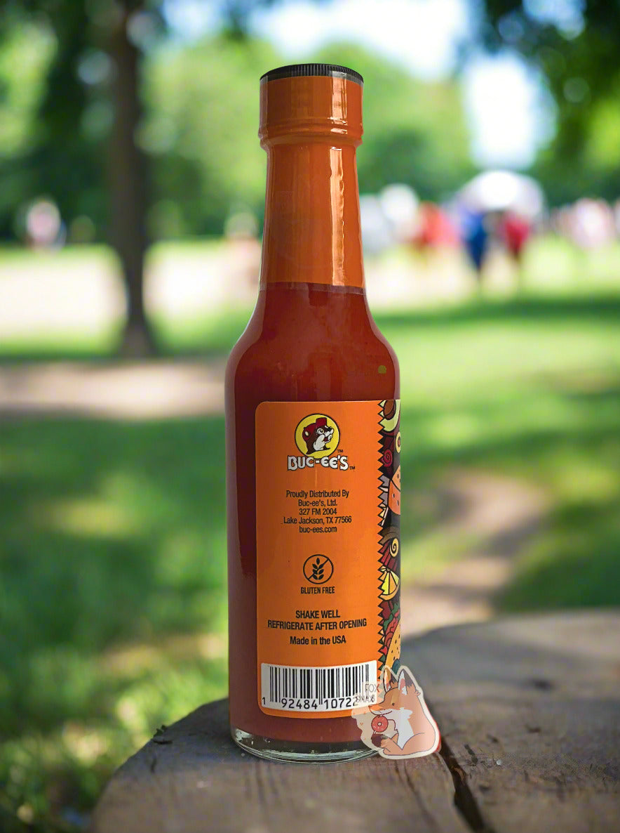 A picture of a small, tall glass bottle of hot sauce. The top of the bottle is sealed with an orange plastic top. The back reads GLUTEN FREE, then SHAKE WELL and REFRIGERATE AFTER OPENING. Made in the USA. The sauce inside is orange/red. The background is of a park on a summer day; the bottle rests on a wooden picnic table.