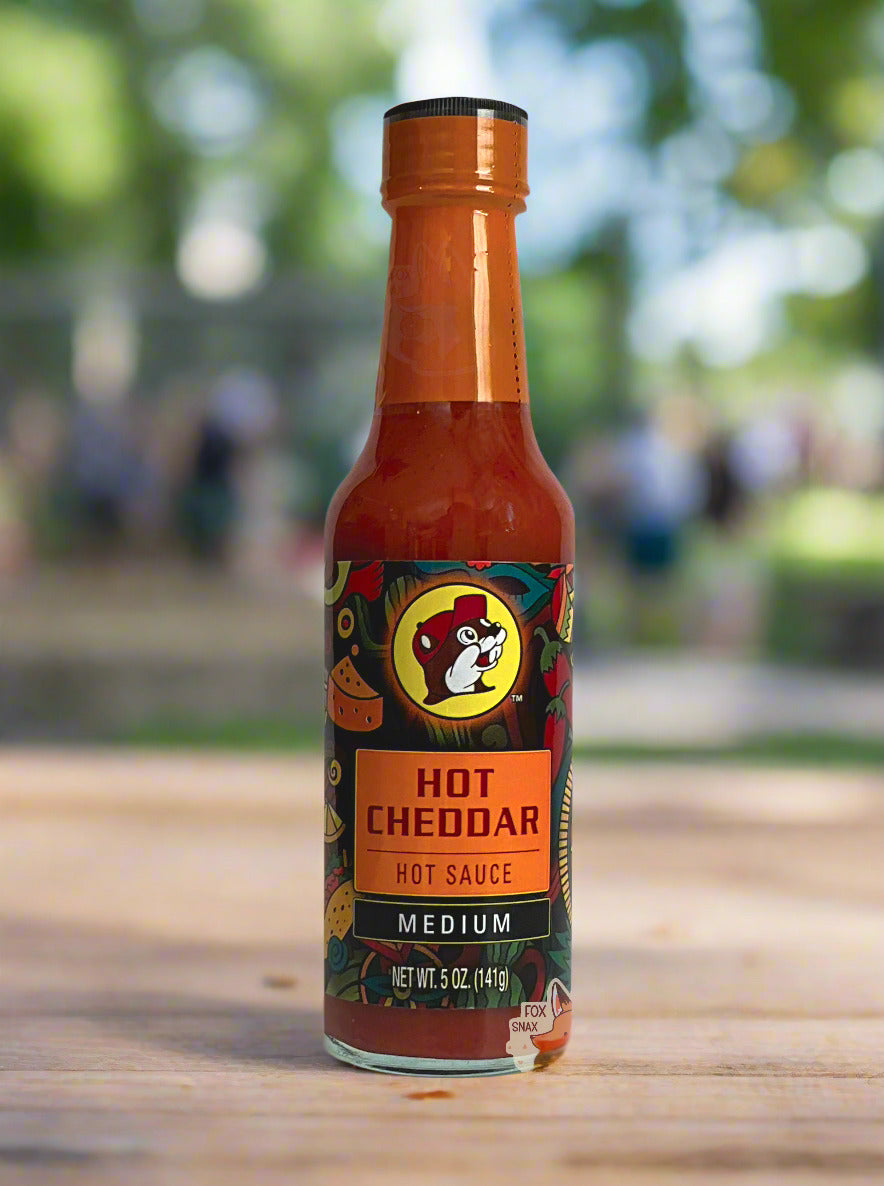A picture of a small, tall glass bottle of hot sauce. The top of the bottle is sealed with a orange plastic cover on a black cap. The front label of the bottle reads HOT CHEDDAR, HOT SAUCE, MEDIUMN. The Buc-ee's logo can be seen on the front; the background is a fairly busy abstract artwork featuring chilis, maracas, aztec wheels, and cheese wedges. The sauce inside is a orange/red. The background is at a park on a hot summer day; multiple people can be seen in the background as well.