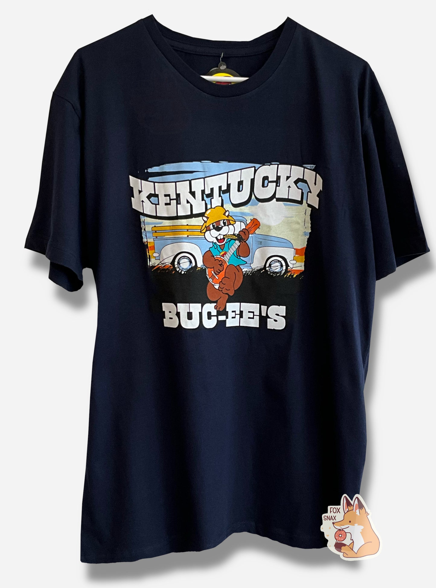 A picture of a dark blue t-shirt, framed on the top and bottom in a white, blocky, print "KENTUCKY", and "BUC-EE'S". In the middle is a beautiful sunset, with Buc-ee's front and center. He's wearing a straw hat (his brown ears poke out of it), a blue/green shirt, and is holding an orange banjo in both hands. A yellow hayseed pokes out of the left side of his mouth, and he's smiling. Behind him is an old-school blue/white pickup truck, framed by tall grass.