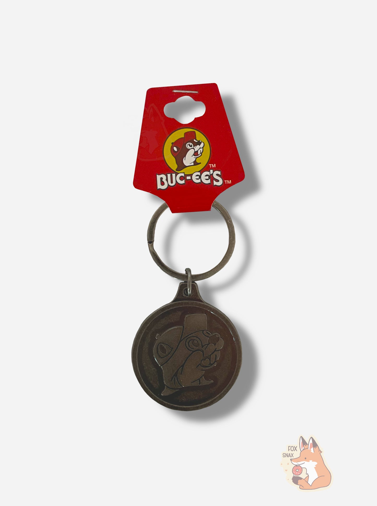A picture of a small dark brownish metal keychain. The keychain is circular and has the Buc-ee the Beaver logo in the middle: a cheerful beaver with a significant overbite, wearing a ball cap. The top of the chain has a small overhang that is mounted to a circular chain for holding keys. The top of the chain has a red paper tag that reads BUC-EE'S.