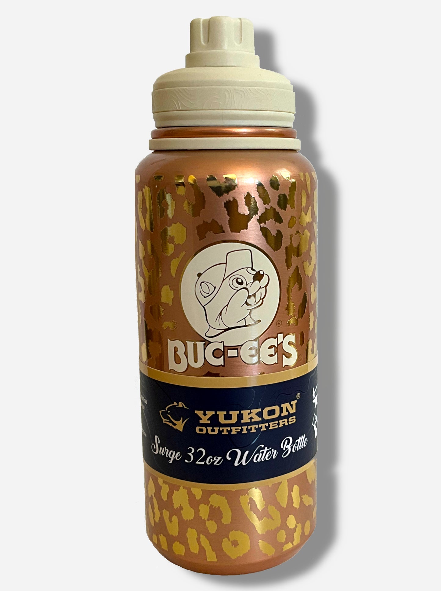 A picture of a tall, metallic, gold leopard print cylindrical water bottle, with a big picture of Buc-ee The Beaver on it. The surface is spotted like a leopard's hide, alternately dull and bright gold, as it is covering stainless steel. There is no handle. The top is a screw on plastic knob with a keychain handle. A paper wrapper around the bottom reads: Yukon Outfitters. Surge 32 oz Water Bottle.