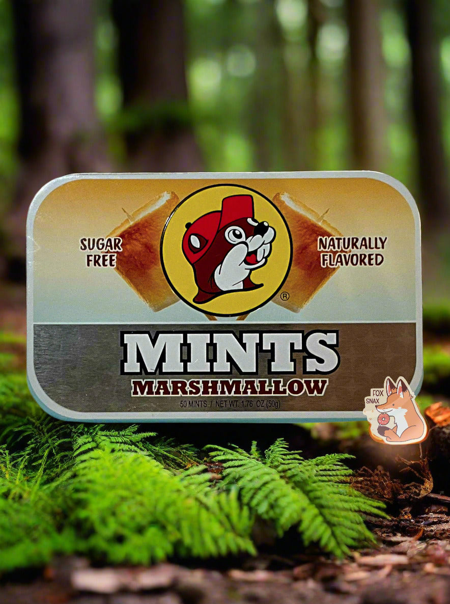 A picture of a tin of Buc-ees Marshmallow Mints. The cover is decorated with a big picture of Buc-ee The Beaver, and has a pair of toasted marshmallows against a tan and silver background. The tin reads Sugar Free, Naturally Flavored, Mints, Marshmallow. The mints are sitting in a pine forest; green ferns frame the bottom of the box of mints, and a campfire can be seen in the right of the picture.
