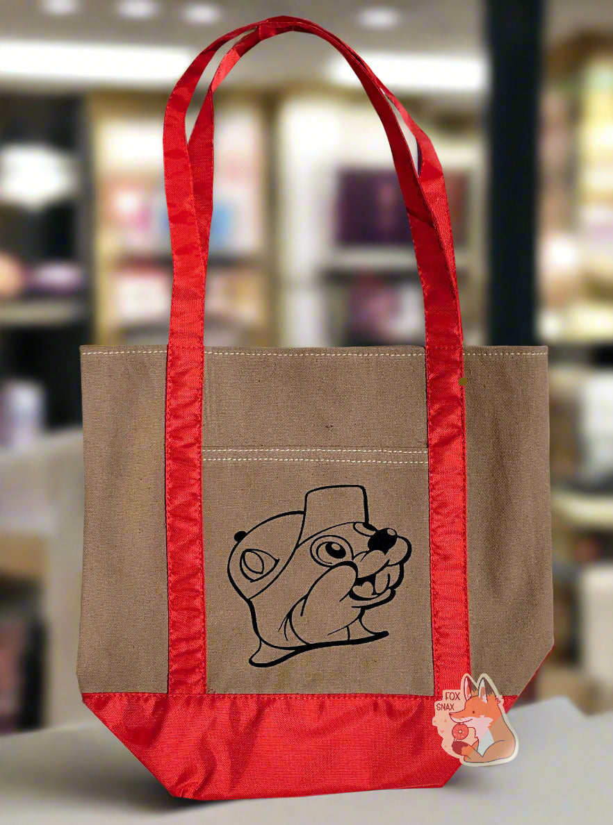 Picture of a small tote bag, with shiny red cloth straps. In the center of the bag is a black outline of Buc-ee The Beaver: a jolly beaver wearing a ballcap and a buc-toothed smile. The bag is sitting on a perfume counter in an upscale mall department store.