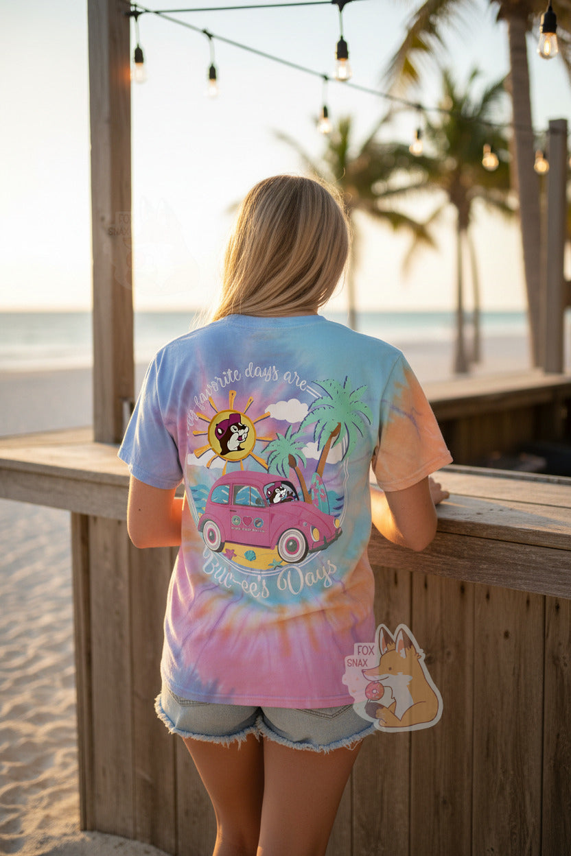 A photo of a woman with long blond hair wearing a multicolor tie-dye shirt. The back of the shirt has a spiral of color; orange, blue, light blue,yellow, pink. In the middle of the chest are is picture of Buc-ee the Beaver wearing a dark pink ballcap. The woman is also wearing jean shorts, and is standing by a beach bar, on a beach at sunset. She is standing with her back towards the viewer. Multiple palm trees can be seen in the distance.