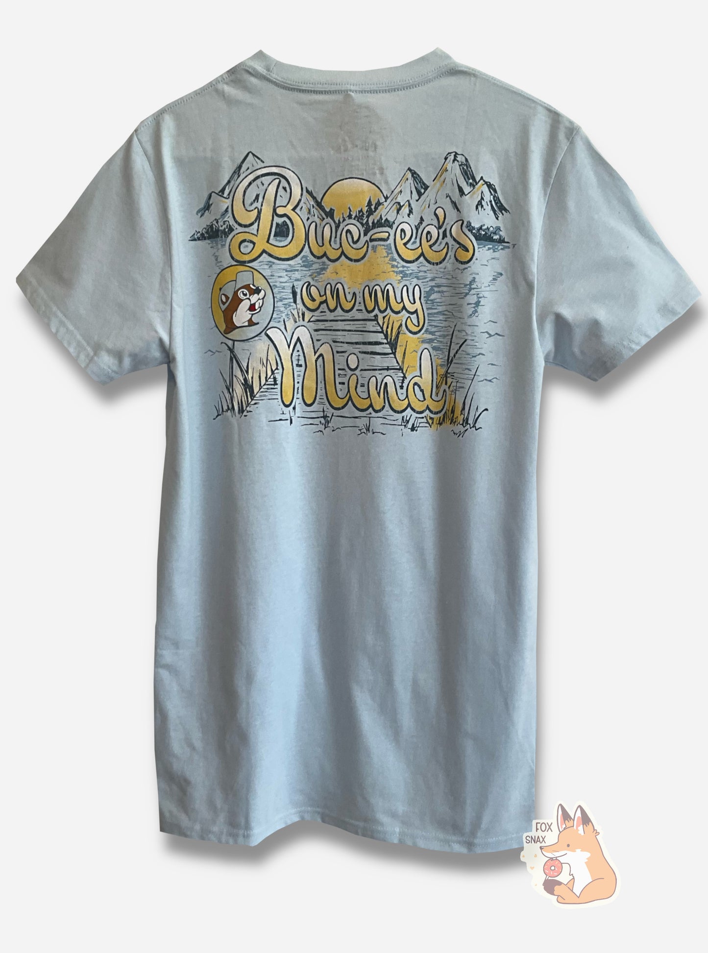 A picture of the back of a a light blue t-shirt. The t-shirt has a drawing of a peaked mountain range, in dark blue, framed on the bottom by calm water and trees. The near side of the water is split in the middle by a large wooden dock framed with tall grasses.
The entire scene is overlaid with cursive yellow text that reads: BUC-EE'S ON MY MIND. On the left of the text is the Buc-ee's logo in a light gradient from yellow to blue. Buc-ee is wearing a light blue ball cap, same as the front of the shirt.