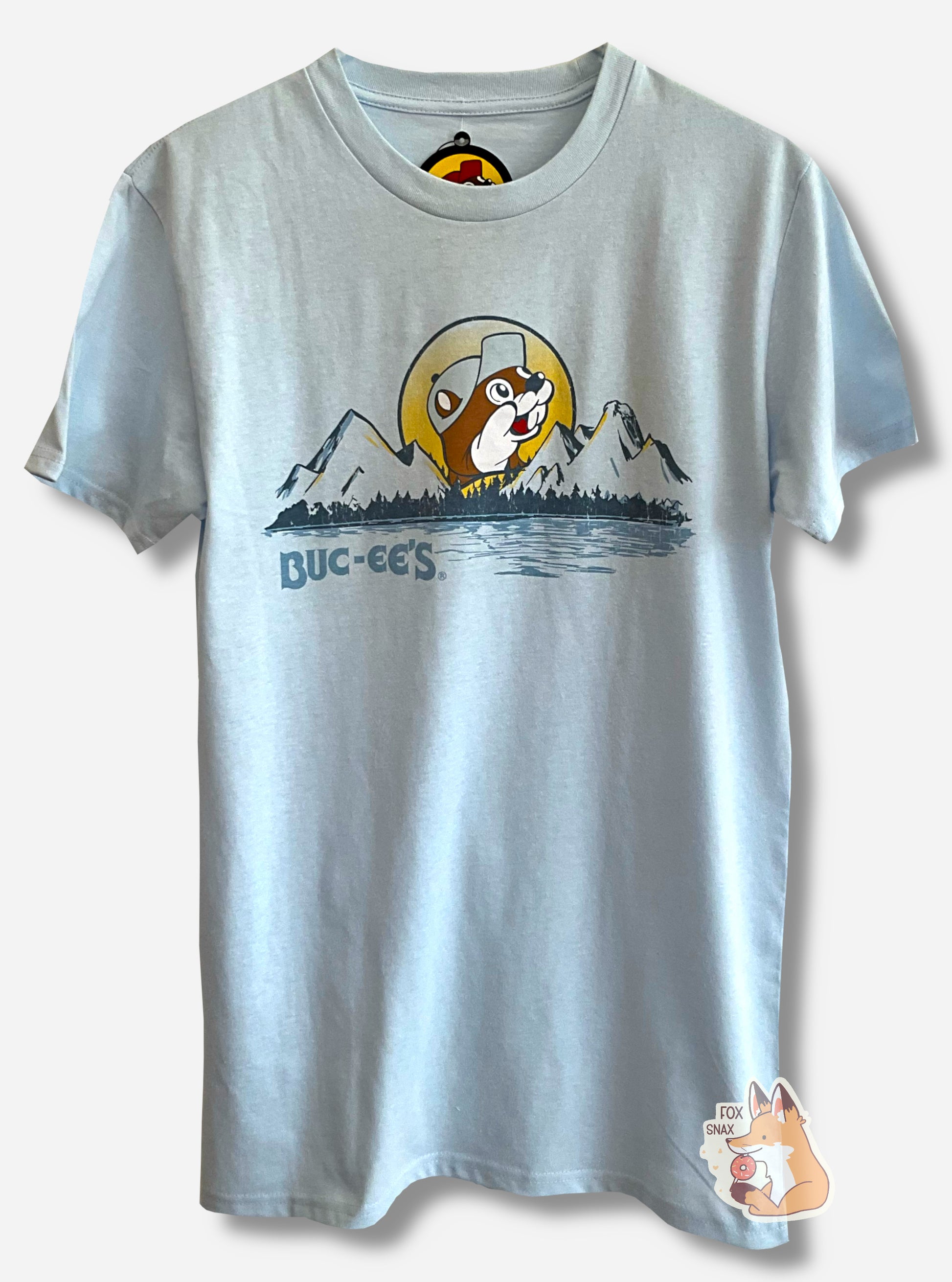 A picture of the front of a light blue t-shirt.  The t-shirt has a drawing of a peaked mountain range, in dark blue, framed on the bottom by calm water and trees.  On the horizon is the setting sun in yellow.  The middle of the sun is the Buc-ee's logo, with the background in yellow, and Buc-ee The Beaver, wearing a light blue cap, inside the sun.  In the lower left of the picture are the words: BUC-EE'S.