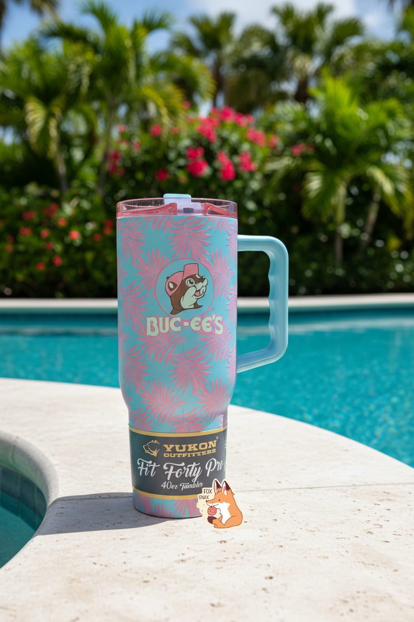 A picture of a tall, light blue, sorta skinny tumbler, with a big picture of Buc-ee The Beaver on it. Buc-ee's hat, and the circle behind him, is in light pink. The surface is light blue, and covered in light pink palm fronds. The handle and sipper on top of the lid are light blue. The top is light transparent pink plastic. A paper wrapper around the bottom reads: Yukon Outfitters. Fit Forty Pro 40 oz Tumbler. The tumbler is sitting on concrete next to a tropical pool.