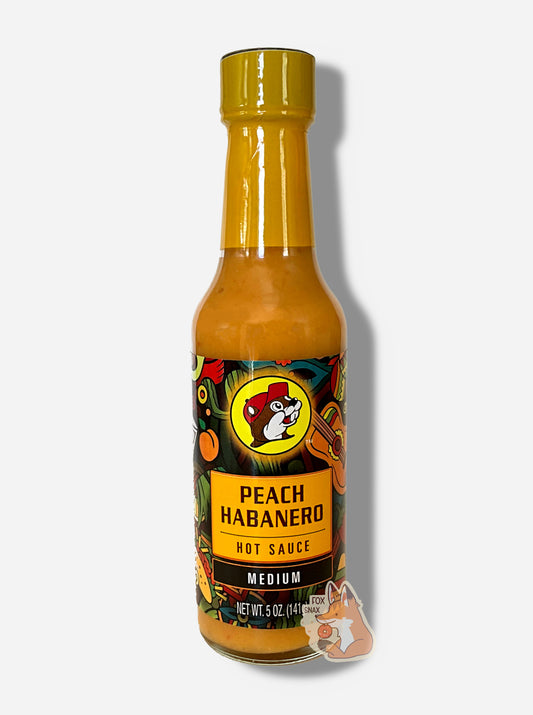 A picture of a small, tall glass bottle of hot sauce.  The top of the bottle is sealed with a orange-yellow plastic cover on a black cap.  The front label of the bottle reads PEACH HABANERO HOT SAUCE, FIERY HOT.  The Buc-ee's logo can be seen on the front; the background is a fairly busy abstract artwork featuring a guitar, a peach, and a taco.  The sauce inside is bright orange / yellow.