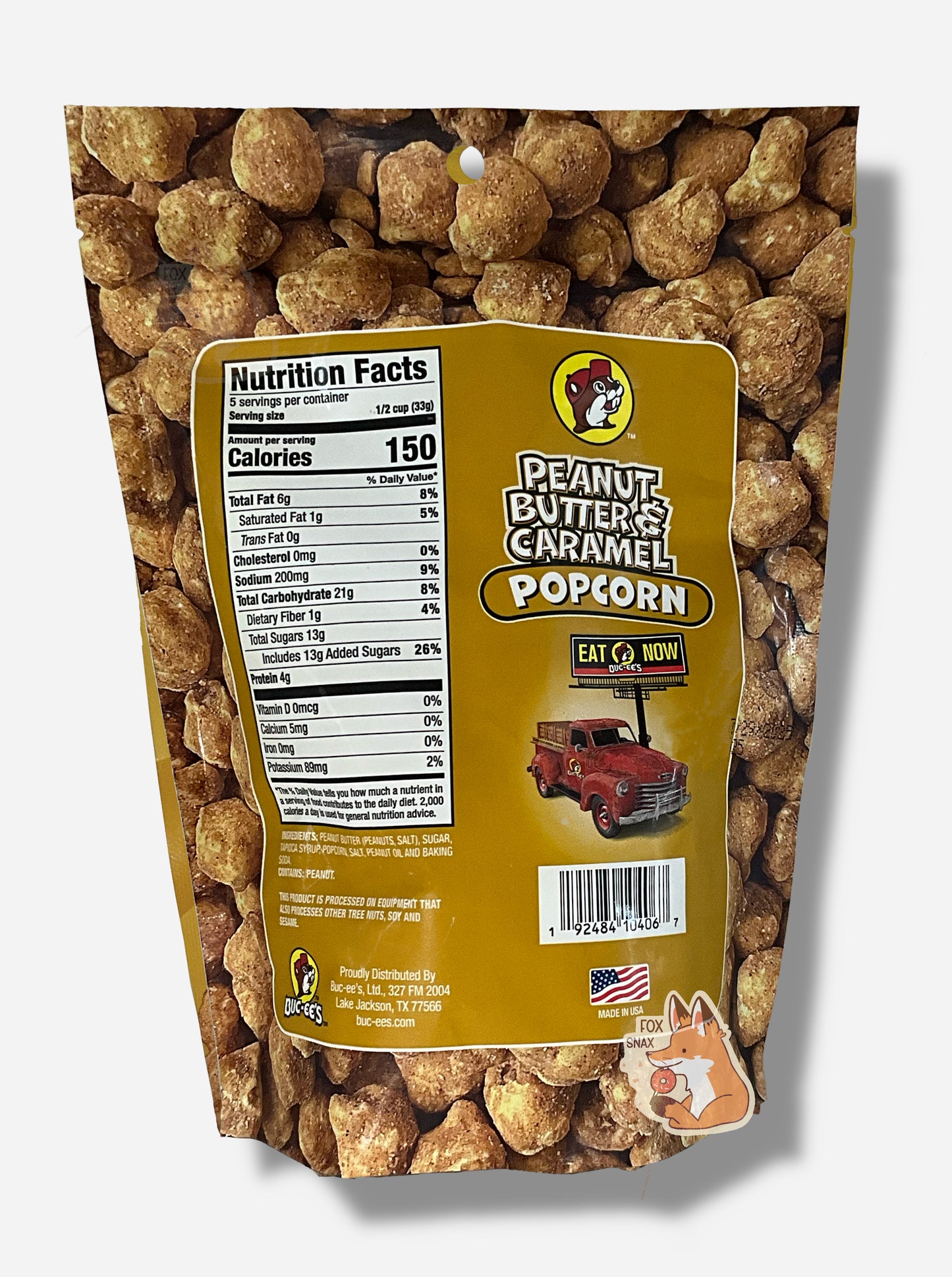 A picture of a tan/yellow bag of Buc-ee's Peanut Butter & Caramel Popcorn. The background is almost solely popcorn nuggets, each coated in brown peanut butter and caramel coating. The right side reads PEANUT BUTTER & CARAMEL POPCORN and has a picture of a red truck under a Buc-ee's EAT NOW sign. Underneath that is the American flag, and says MADE IN USA.