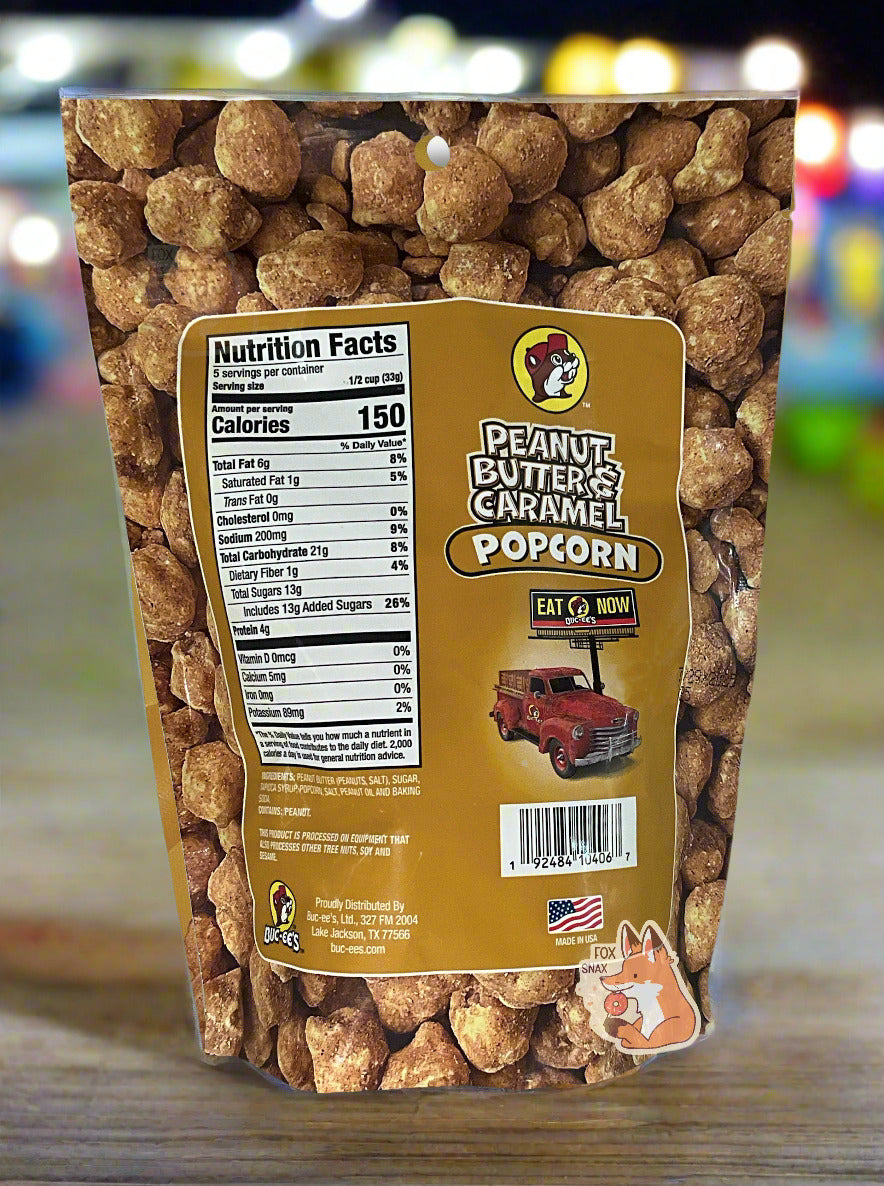 A picture of a tan/yellow bag of Buc-ee's Peanut Butter & Caramel Popcorn. The background is almost solely popcorn nuggets, each coated in brown peanut butter and caramel coating. The right side reads PEANUT BUTTER & CARAMEL POPCORN and has a picture of a red truck under a Buc-ee's EAT NOW sign. Underneath that is the American flag, and says MADE IN USA. The bag is sitting on a wooden bench; in the background, the bright lights of a county fair can be seen.