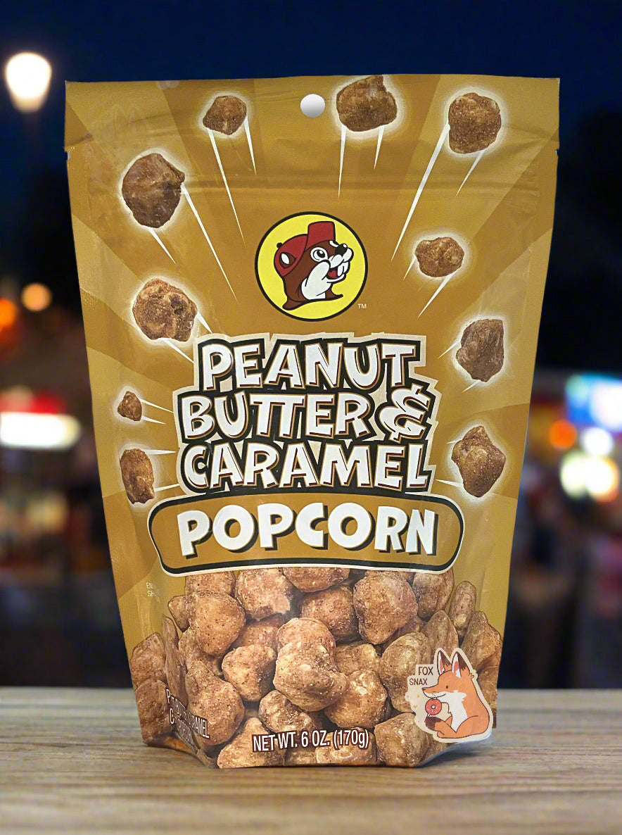 A picture of a tan/yellow bag of Buc-ee's Peanut Butter & Caramel Popcorn. The front of the bag has a group of popcorn nuggets, each coated in brown peanut butter and caramel coating, exploding from the center. A pile of popcorn is on the bottom. The top center has the Buc-ee's logo, with cheerful beaver wearing a red ball cap, framed in a yellow circle, and reads PEANUT BUTTER & CARAMEL POPCORN. The bag is sitting on a wooden bench; in the background, the bright lights of a county fair can be seen.