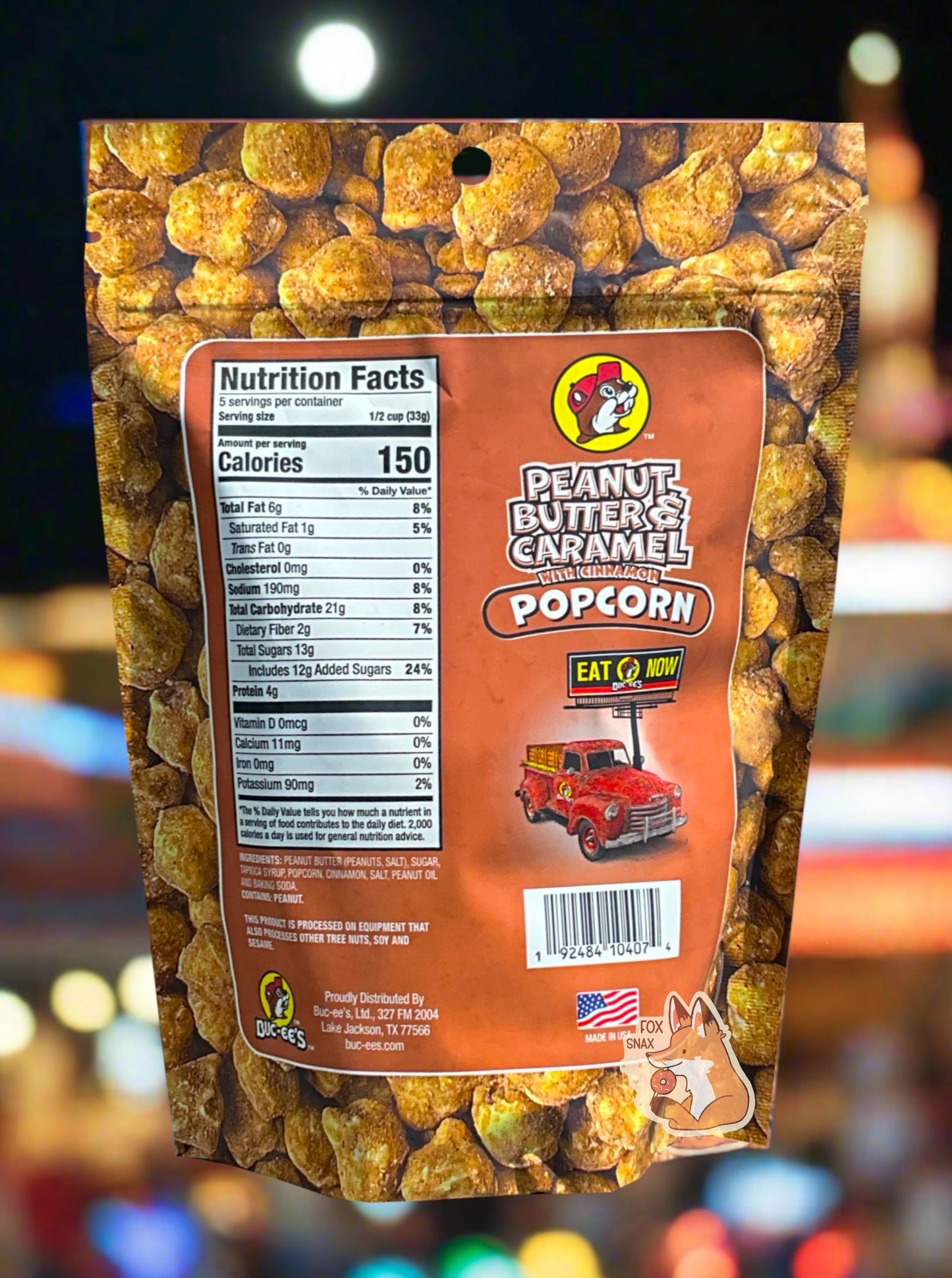 A picture of a red/pink bag of Buc-ee's Peanut Butter & Caramel Popcorn. The background is almost solely popcorn nuggets, each coated in brown peanut butter, cinnamon, and caramel coating. The right side reads PEANUT BUTTER & CARAMEL WITH CINNAMON POPCORN and has a picture of a red truck under a Buc-ee's EAT NOW sign. Underneath that is the American flag, and says MADE IN USA. The background shows a county fair, complete with multicolored lights.