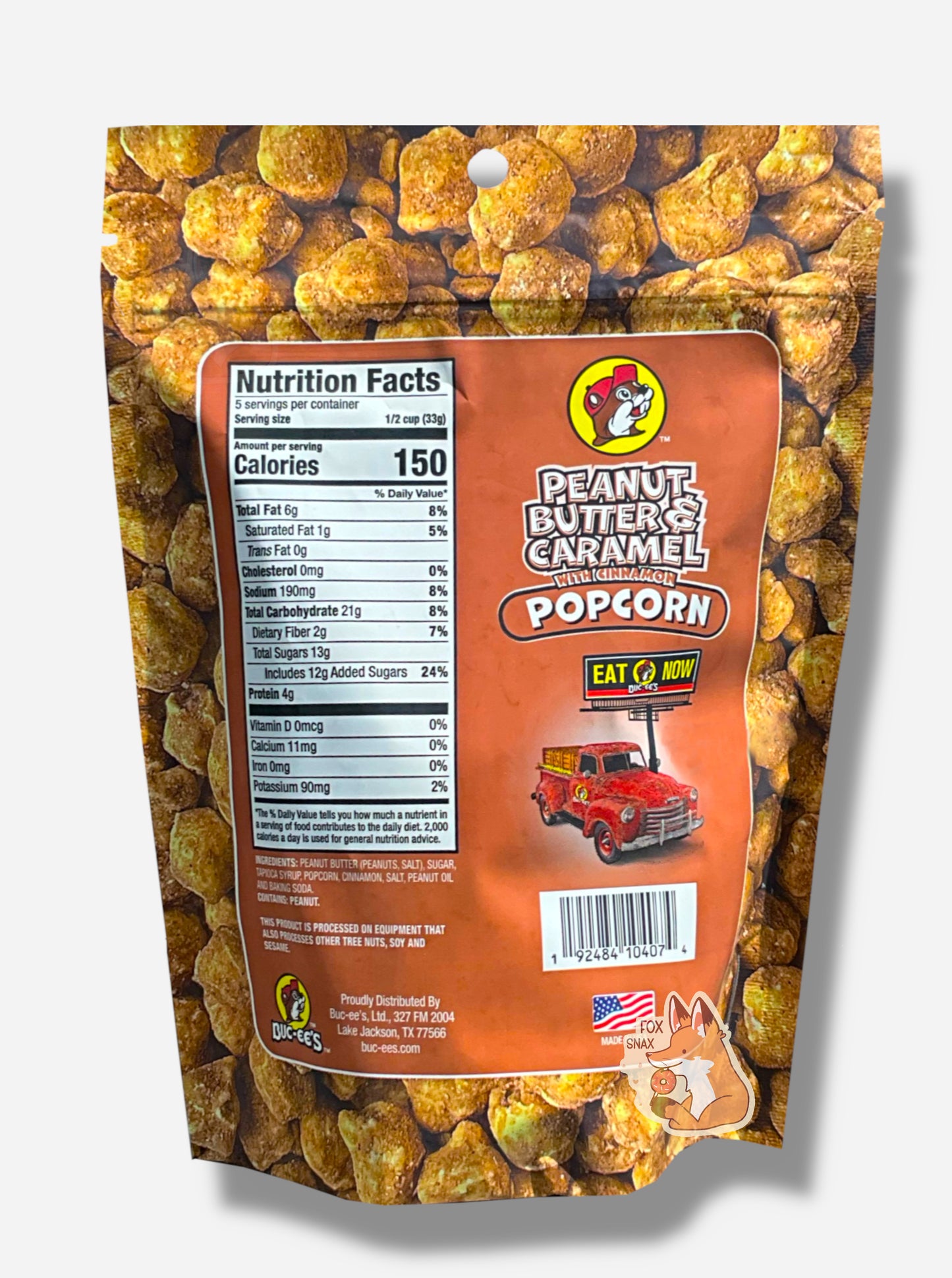 A picture of a red/pink bag of Buc-ee's Peanut Butter & Caramel Popcorn. The background is almost solely popcorn nuggets, each coated in brown peanut butter, cinnamon, and caramel coating. The right side reads PEANUT BUTTER & CARAMEL WITH CINNAMON POPCORN and has a picture of a red truck under a Buc-ee's EAT NOW sign. Underneath that is the American flag, and says MADE IN USA.