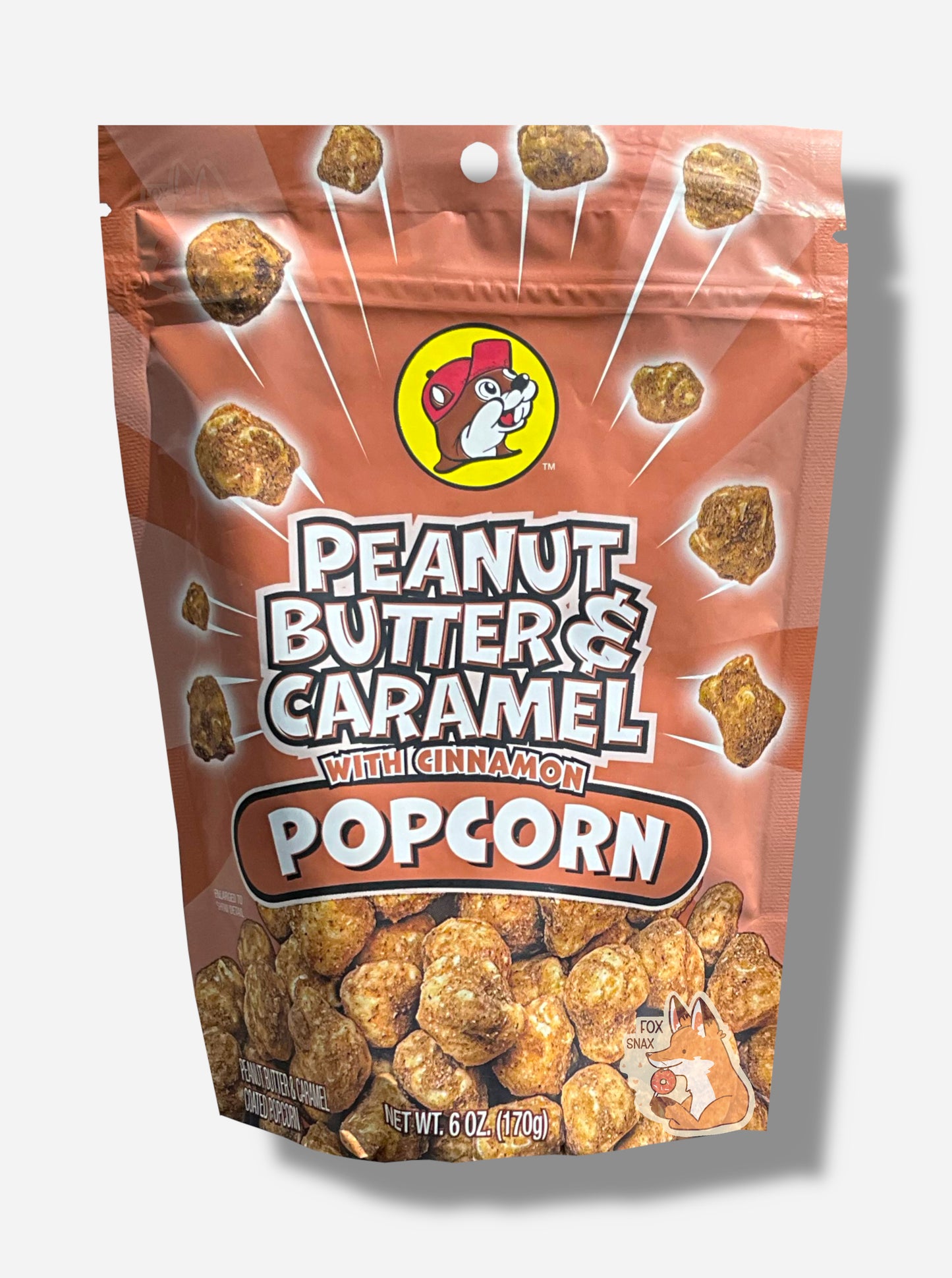 A picture of a red/pink bag of Buc-ee's Peanut Butter & Caramel Popcorn With Cinnamon. The front of the bag has a group of popcorn nuggets, each coated in brown peanut butter, caramel, and cinnamon coating, exploding from the center. A pile of popcorn is on the bottom. The top center has the Buc-ee's logo, with cheerful beaver wearing a red ball cap, framed in a yellow circle, and reads PEANUT BUTTER & CARAMEL WITH CINNAMON POPCORN.