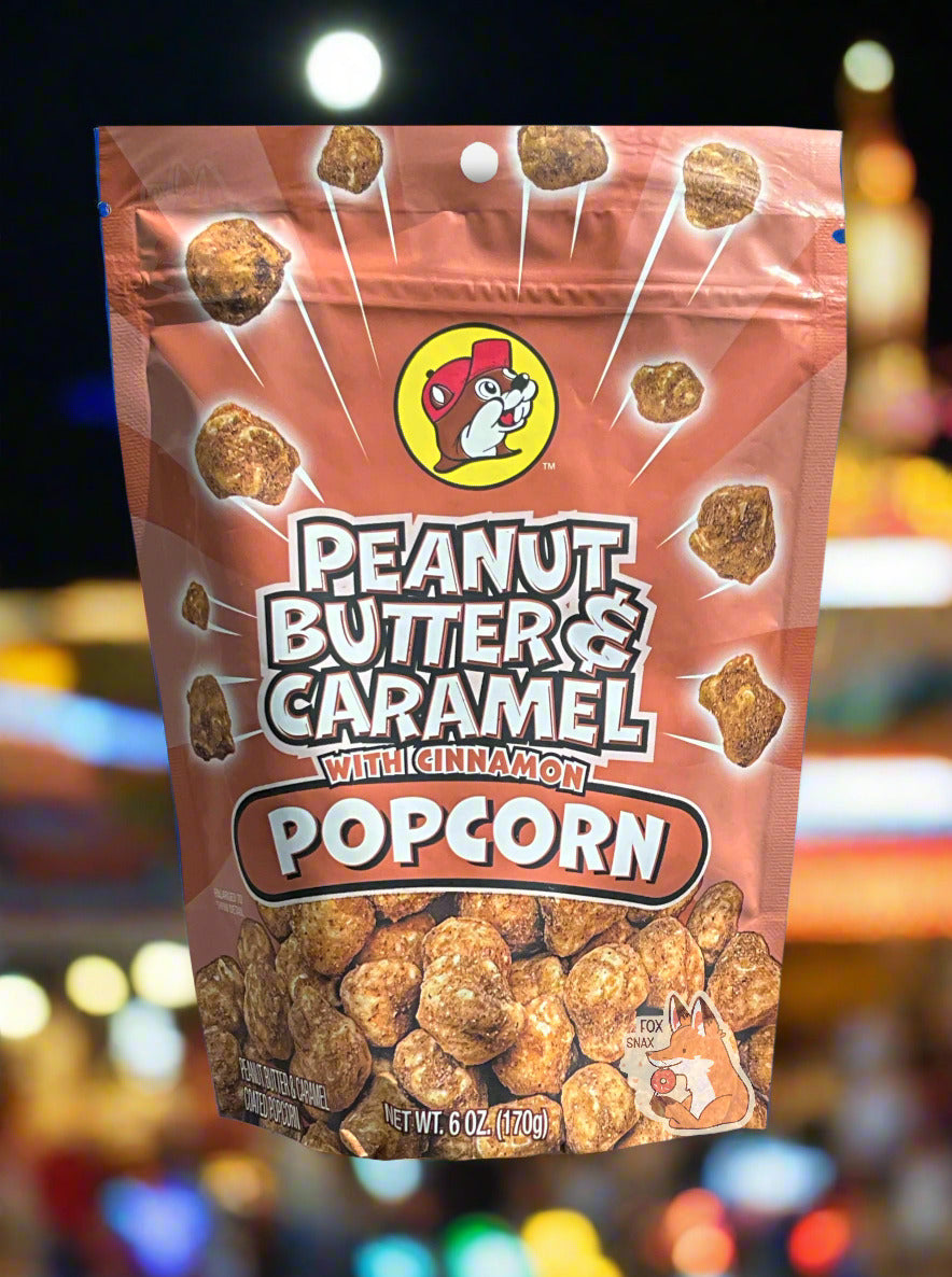 A picture of a red/pink bag of Buc-ee's Peanut Butter & Caramel Popcorn With Cinnamon. The front of the bag has a group of popcorn nuggets, each coated in brown peanut butter, caramel, and cinnamon coating, exploding from the center. A pile of popcorn is on the bottom. The top center has the Buc-ee's logo, with cheerful beaver wearing a red ball cap, framed in a yellow circle, and reads PEANUT BUTTER & CARAMEL WITH CINNAMON POPCORN. The background shows a county fair, complete with multicolored lights.