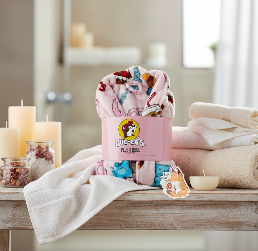 An image of a plush, wrapped, pink robe. The surface is pink and fluffy, and is studded with blue and pink gummi bear pictures, drink cup picture, pretzels, brisket sandwiches, and the text "BUC-EE'S" in red and blue. A cardboard wrapper around the "waist" of the robe shows the Buc-ee's logo and reads "PLUSH ROBE", then, underneath that "ONE SIZE FITS MOST". The wrapped robe is standing upright on a fancy bathroom white-wood table, surrounded by plush towels and candles; in the background is a bathtub.