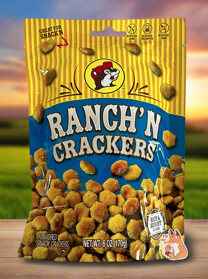 A picture of a metallic bag of Buc-ee's Ranch'N Crackers. The logo is a blue background on white text; a large number of tan, savory-looking hexagonal oyster crackers are shown beneath it. At the top is a picture of the logo of Buc-ee The Beaver, and it reads GREAT FOR SNACK'N, NO ARTIFICIAL COLORS, PRESERVATIVES, FLAVORS. The background is of a meadow, framed against a beautiful multicolored sunset.
