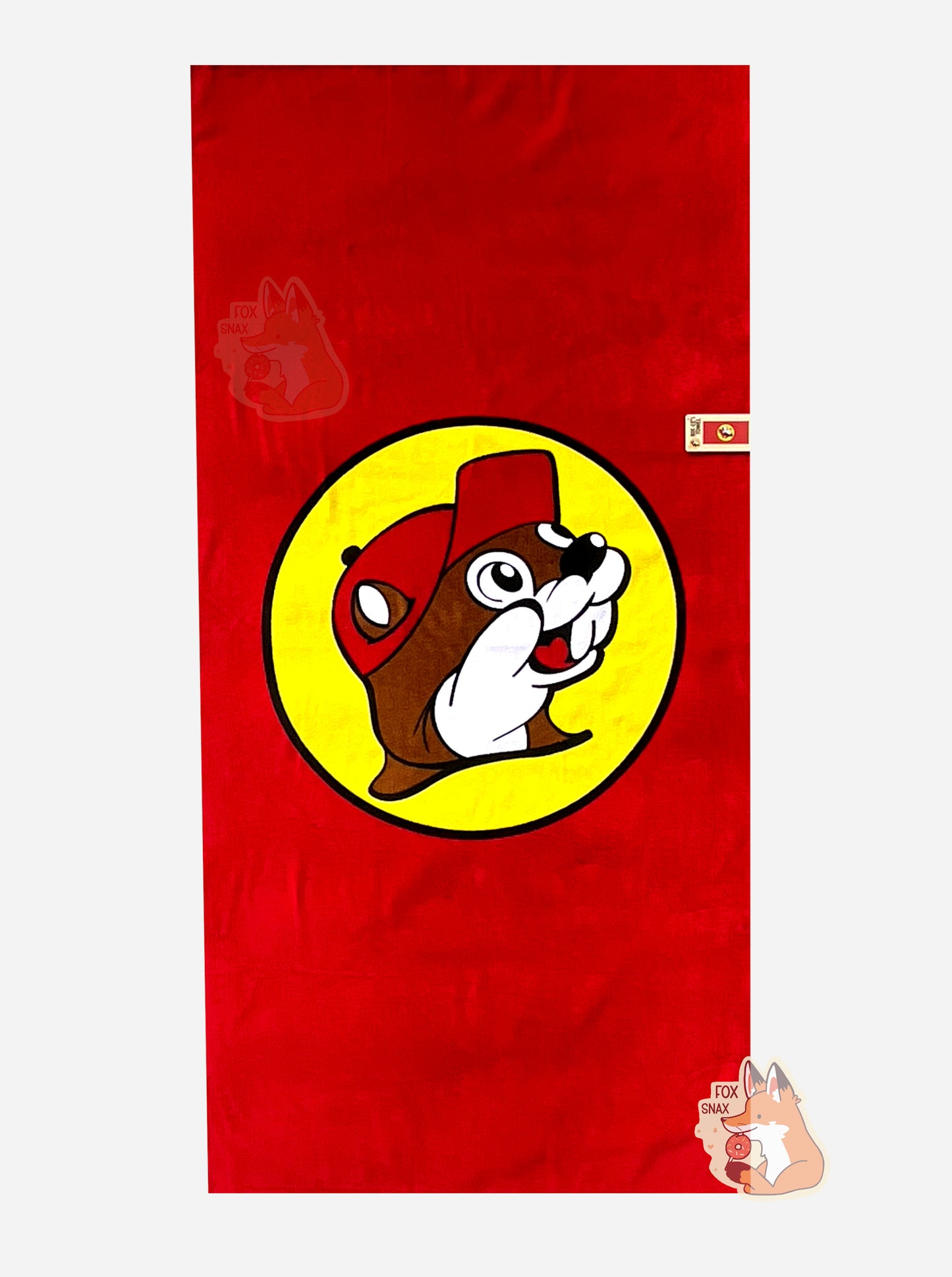 A picture of an unwrapped, 32x64, fluffy beach towel, adorned with a single Buc-ee The Beaver logo. Buc-ee is framed by a yellow circle, and is wearing a red ball cap. A small paper tag on the bottom states: BUC-EE'S TOWEL. 32 in x 64 in. 100% Cotton.