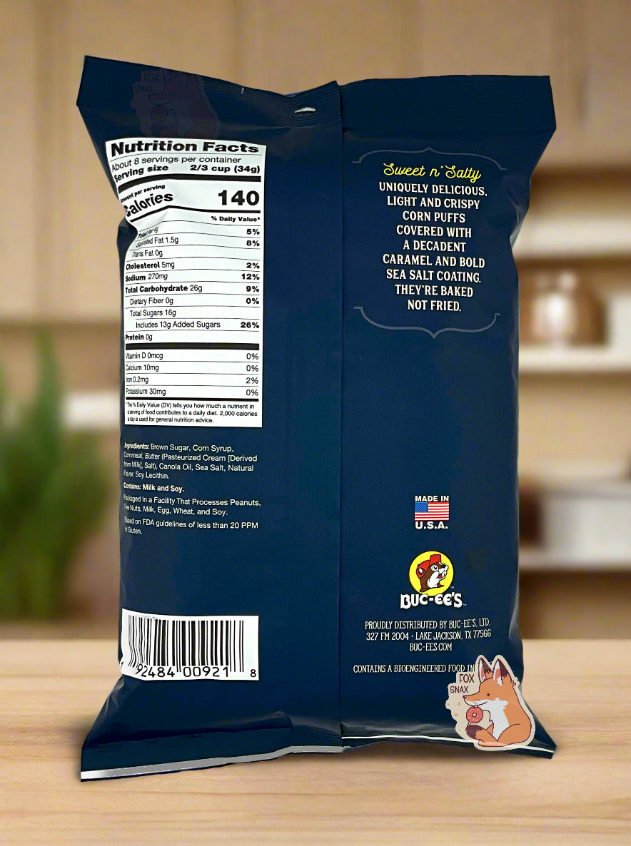 A picture of the back of a opaque, dark blue and white bag of Buc-ee's Sea Salted Caramel Beaver Nuggets. The bag reads: Sweet n' Salty, Uniquely delicious, Light And Crispy, Corn Puffs covered with a decadent caramel and bold sea salt coating. They're baked not fried. The nuggets are sitting on a wooden kitchen countertop.