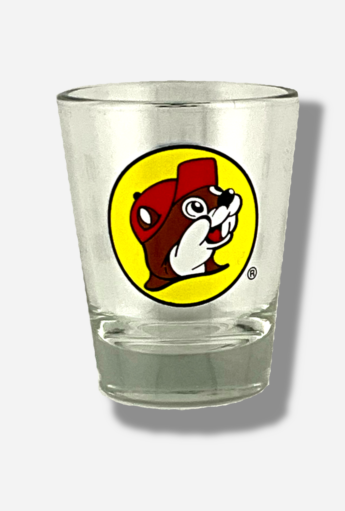 A picture of the front of glass shot glass. The glass is clear. In the middle of the front is the Buc-ee's logo appears inside a yellow circle; it is a cheerful beaver wearing a red ballcap, against a yellow circle background.